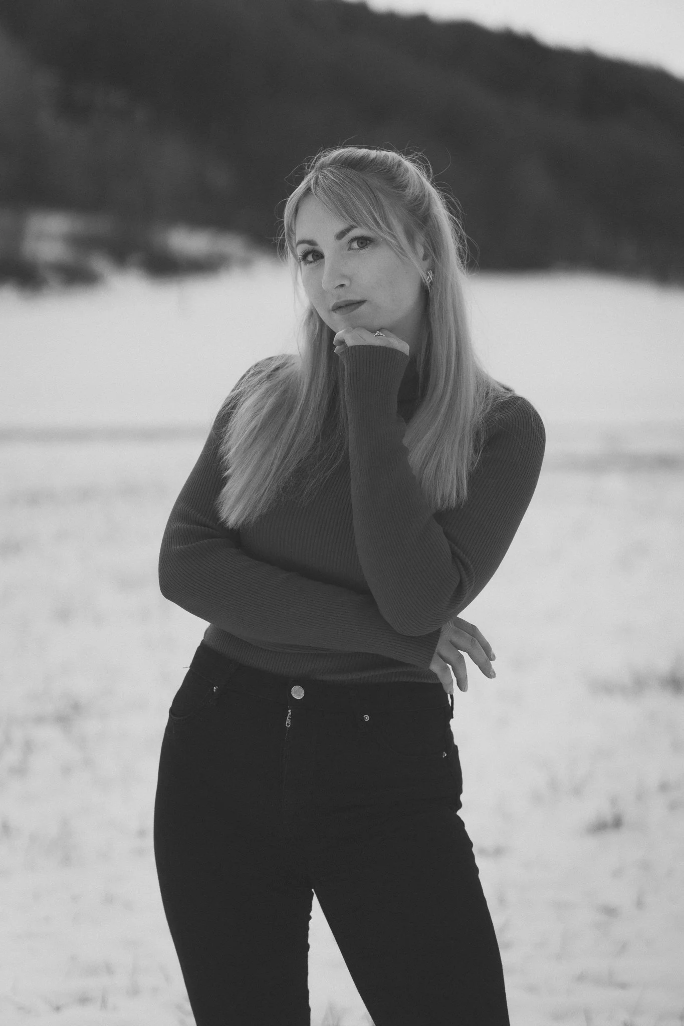 Schwarz-weiß-Foto einer jungen Frau mit langem Haar, die an einem Strand steht, mit einer Hand am Kinn und die andere auf der Hüfte, bei bewölktem Himmel.