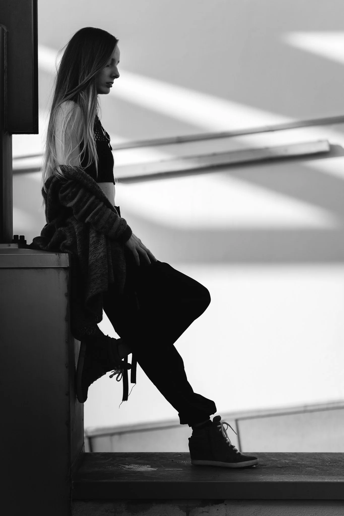 Schwarz-Weiß-Foto einer jungen Frau, die auf einem Platz in einer modernen Architektur sitzt, mit langem Haar, casual Kleidung und Sneakers.