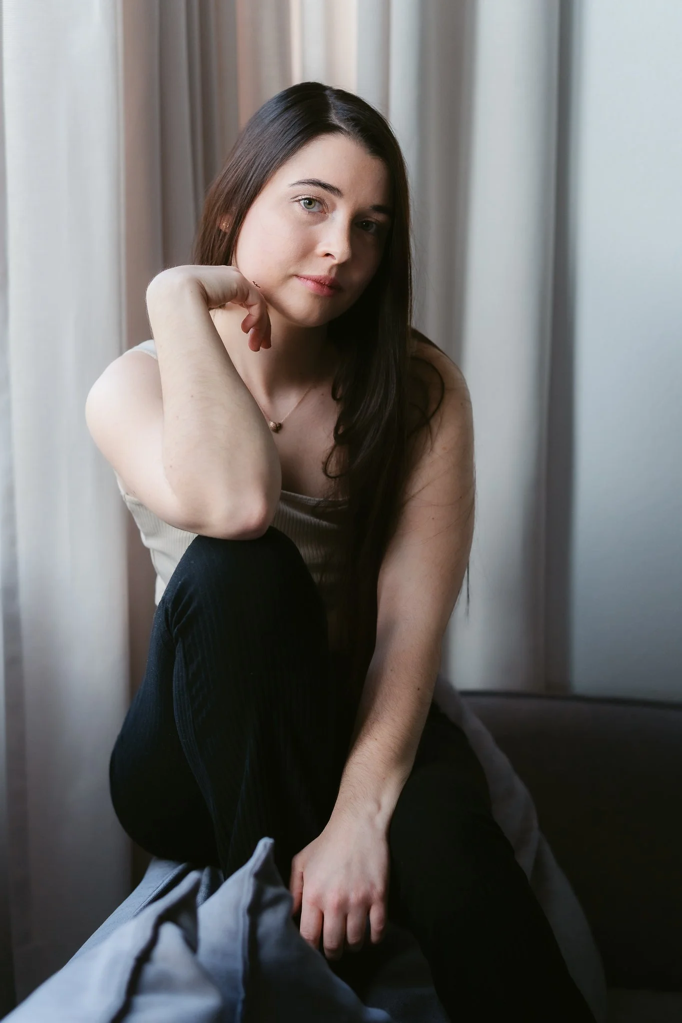 Junge Frau mit langen dunklen Haaren sitzt auf einem Bett vor einem Fenster mit Vorhängen, trägt ein braunes Oberteil und schwarze Hosen, schaut nach vorne.