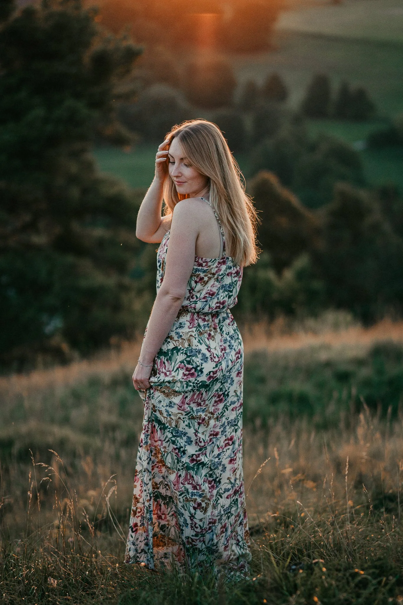 Eine Frau in einem langen, blumigen Kleid steht in einem Feld bei Sonnenuntergang, wirft den Blick leicht nach unten, die Haare sind offen.