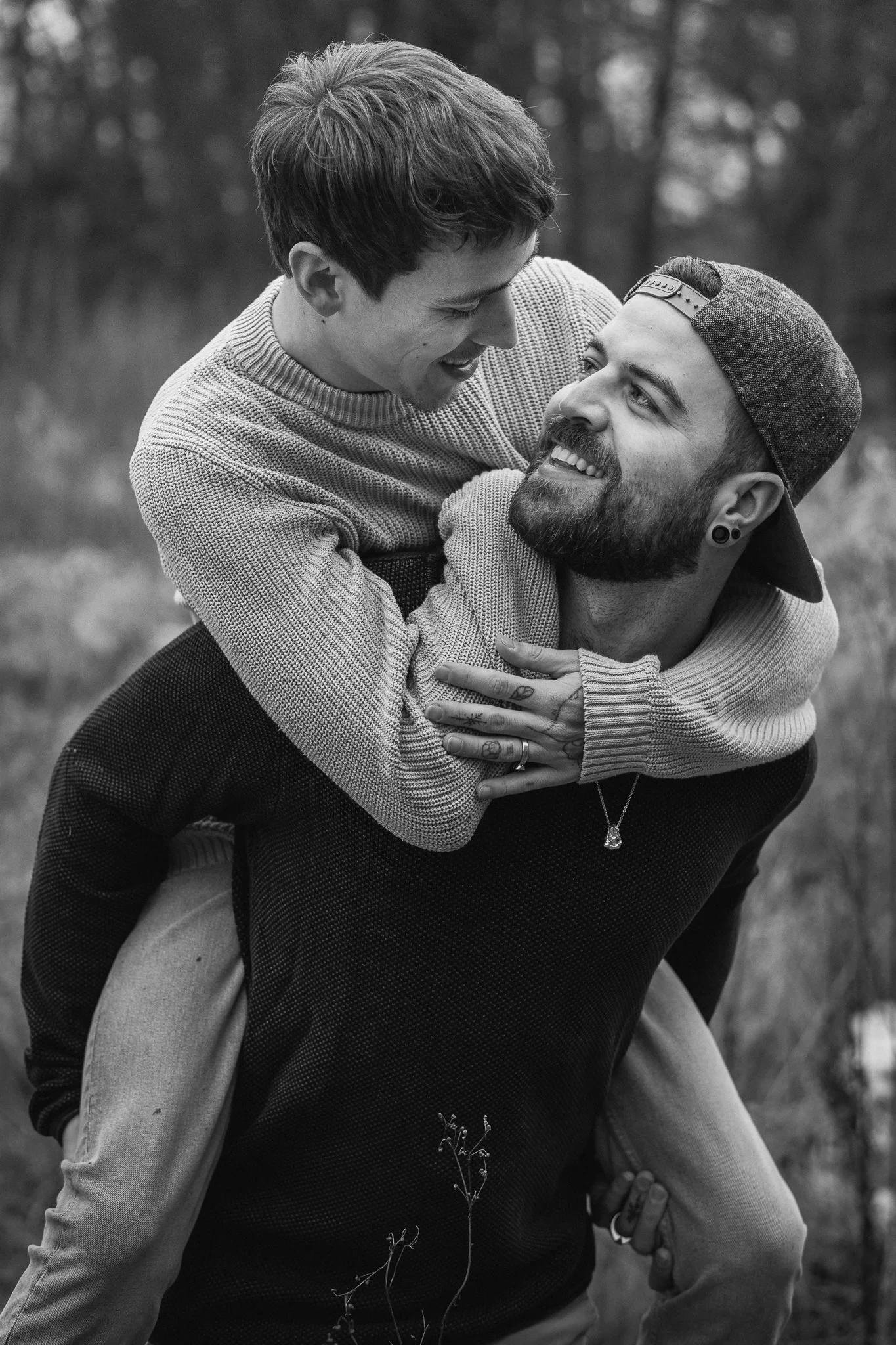 Zwei Männer lachen und umarmen sich im Freien. Einer trägt eine Mütze und hat einen Bart, der andere hat kurze Haare. Es ist eine schwarze und weiße Fotografie.