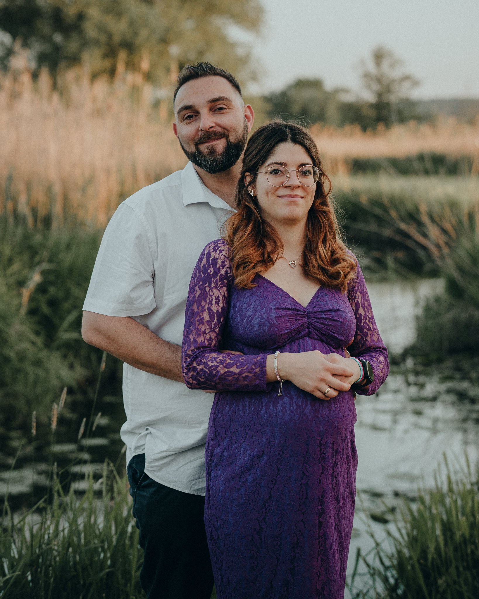 Ein Mann und eine Frau stehen nebeneinander vor einer Wasserlandschaft im Freien, umgeben von Gräsern und Bäumen bei Sonnenuntergang.
