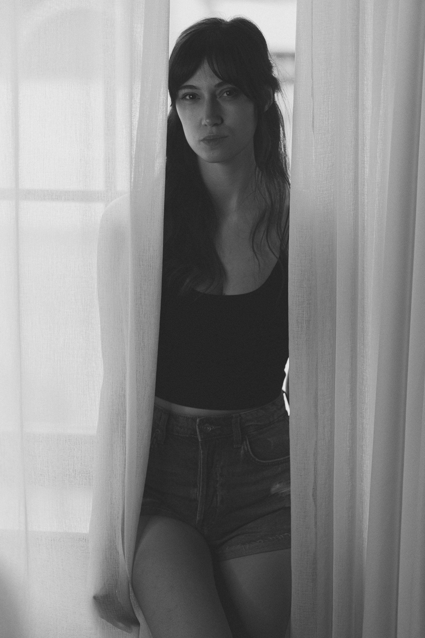 Eine junge Frau blickt durch eine halb geöffneten Vorhänge, trägt ein schwarzes Oberteil und Jeans, die auf einem Stuhl sitzt, im Schwarz-Weiß-Foto.