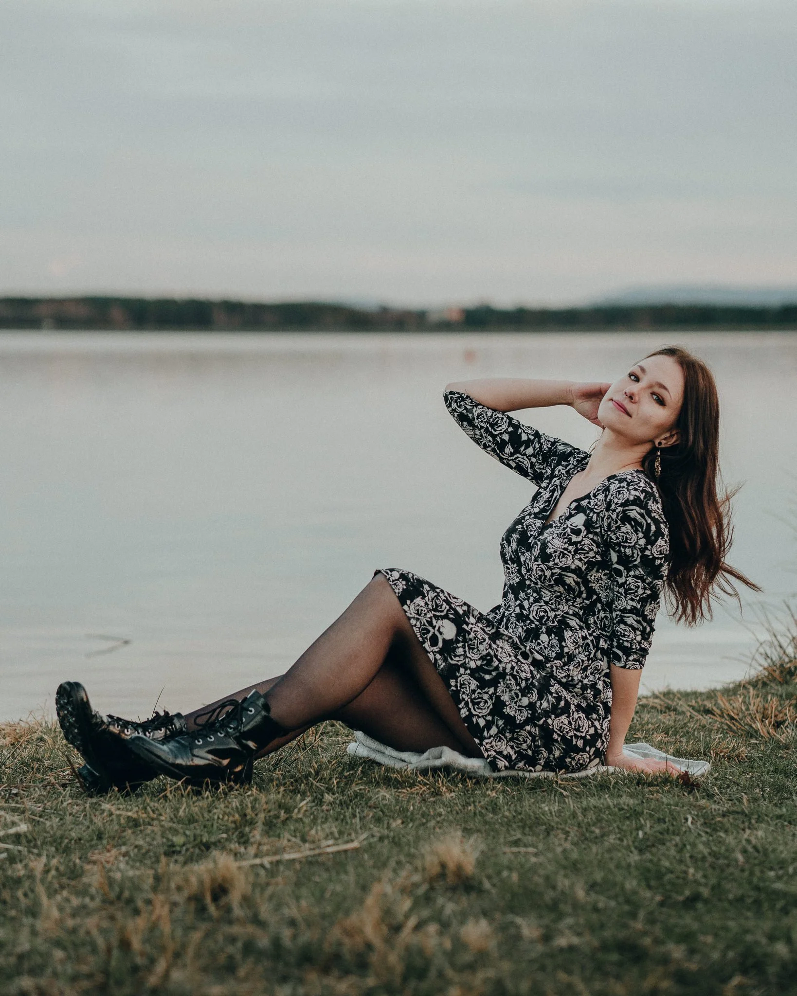 Eine junge Frau sitzt am Ufer eines Sees, trägt ein schwarzes Kleid mit weißen Mustern, schwarze Netzstrümpfe und Stiefel, bei Sonnenuntergang.