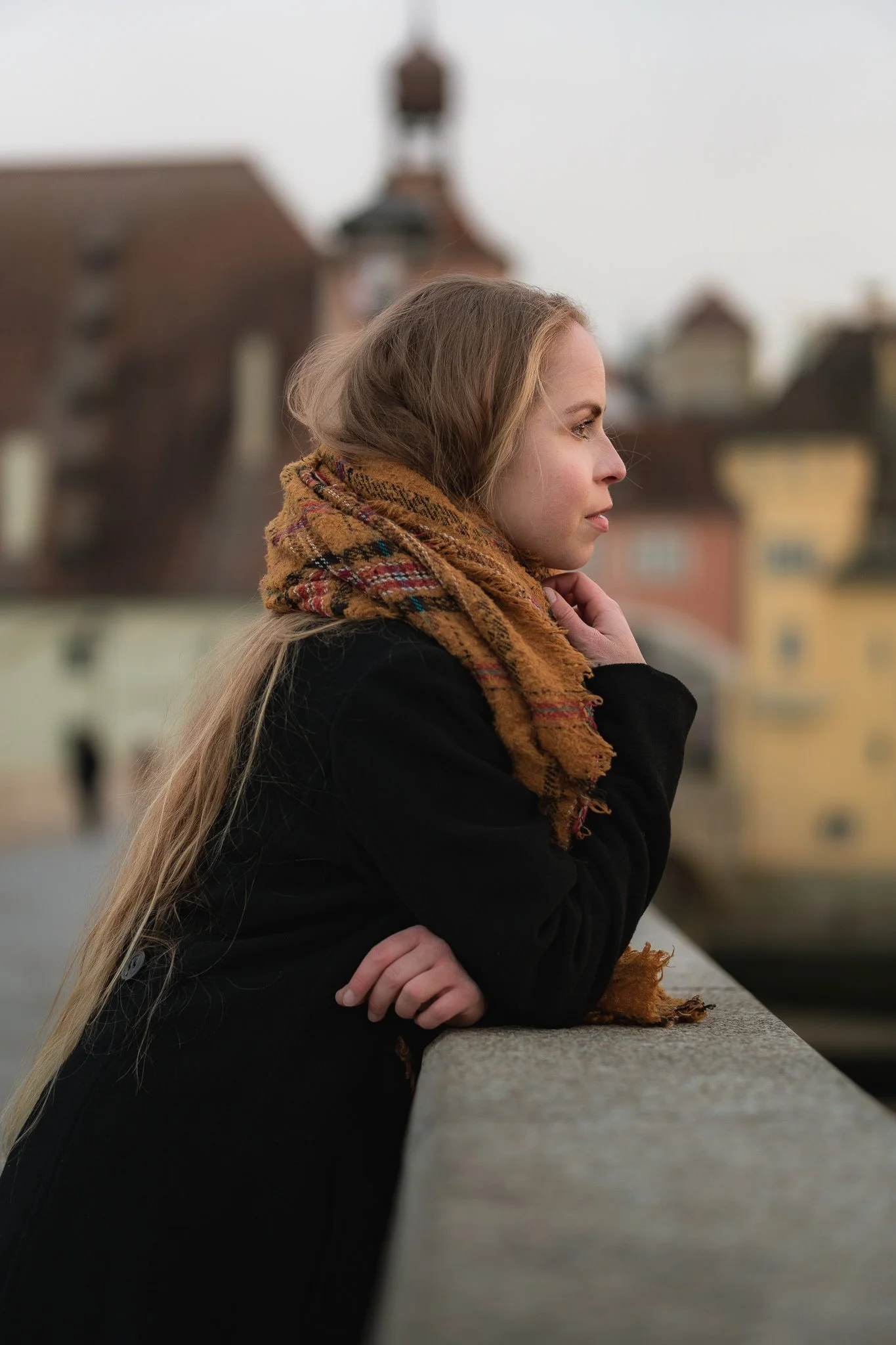 Junge Frau mit langen blonden Haaren, schwarzer Jacke und braun-meliertem Schal lehnt an einem Geländer und schaut nach vorne auf eine Wasserstraße mit bunten Gebäuden im Hintergrund.
