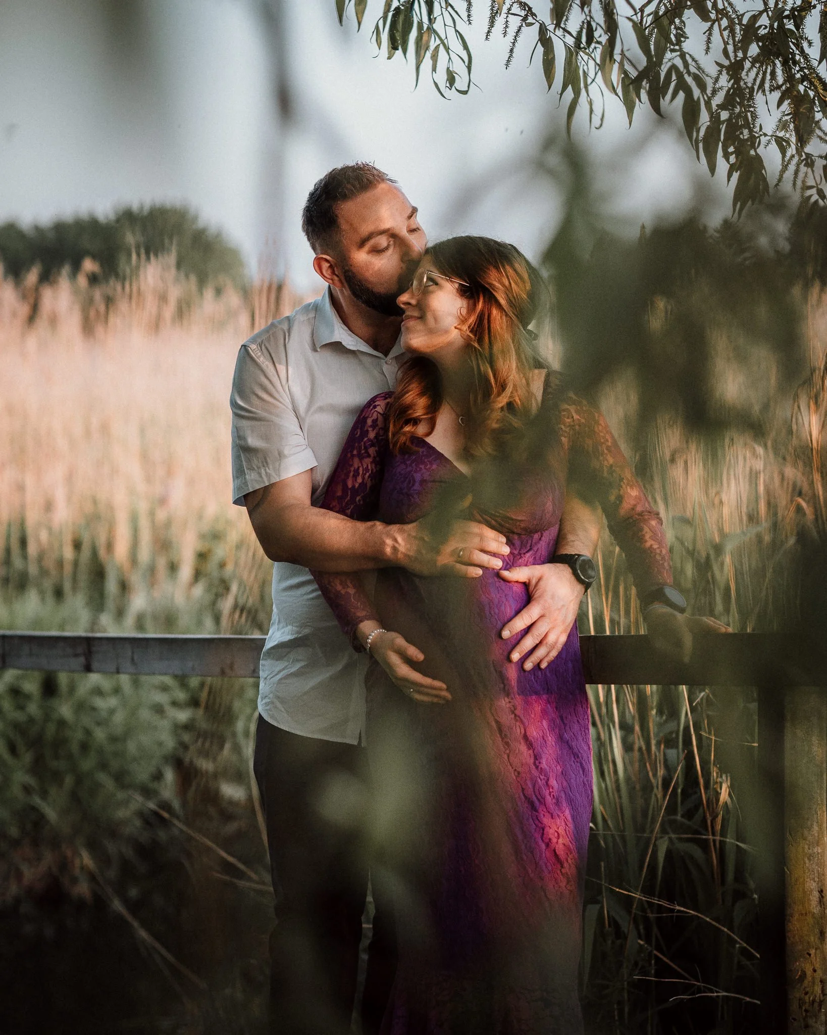 Ein Paar umarmt sich liebevoll im Freien bei Sonnenuntergang, die Frau ist schwanger und trägt ein lila Spitzenkleid, die Umgebung ist grün und natürlich.