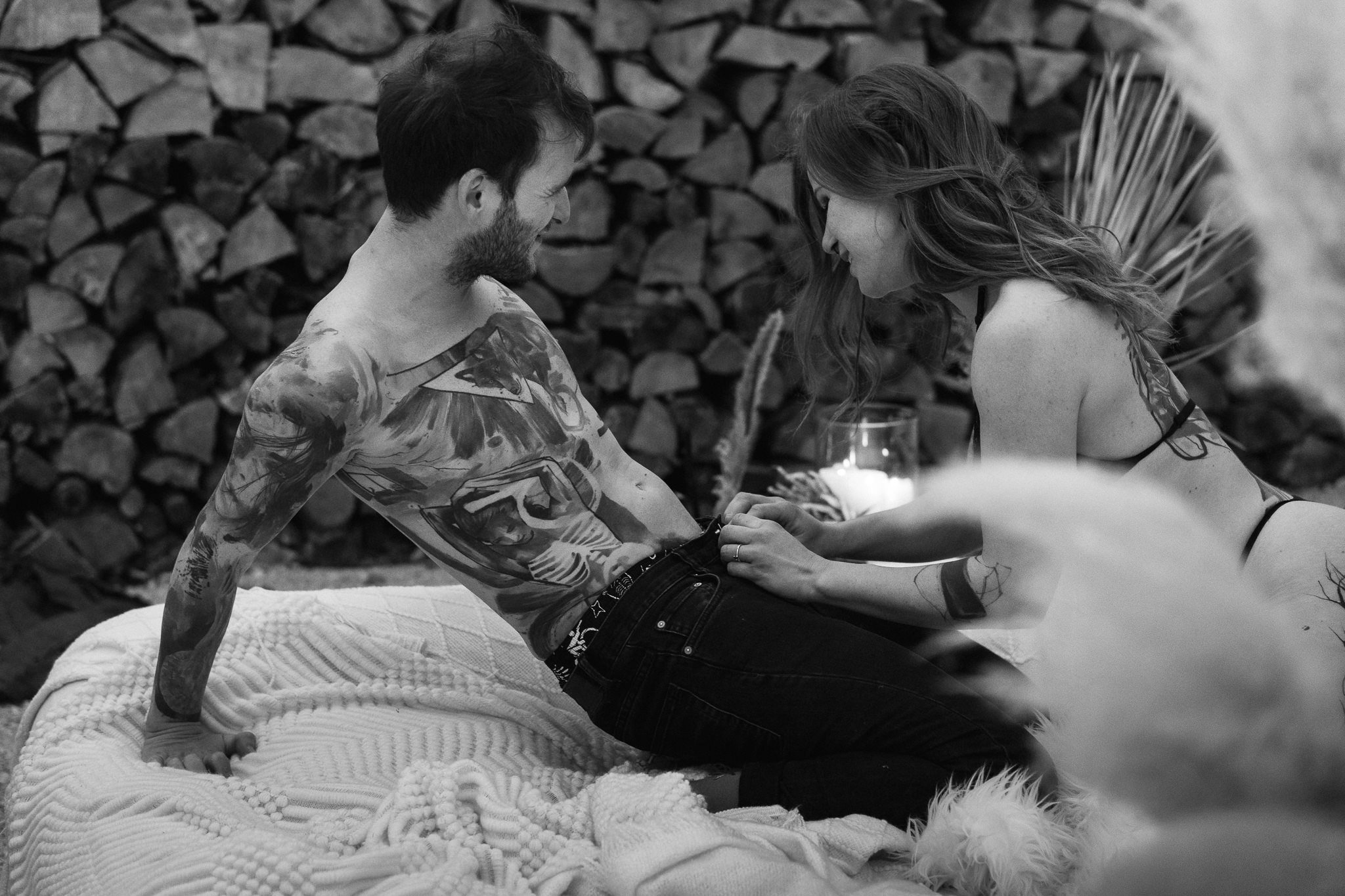 Ein Mann und eine Frau liegen auf einer Decke, die dicht an einem Holzstapelfeld liegt, und schauen sich an. Sie sind halb nackt, die Frau trägt ein Bikini-Oberteil und der Mann trägt keine Oberbekleidung, nur Hosen. Es ist eine intime, liebevolle Sz