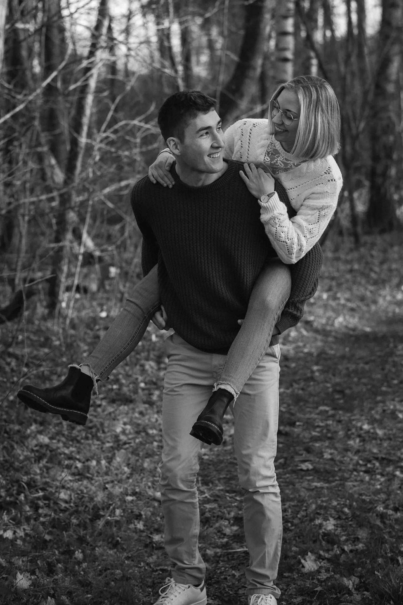 Ein Mann trägt eine Frau auf den Schultern im Wald, beide lachen und haben Spaß.