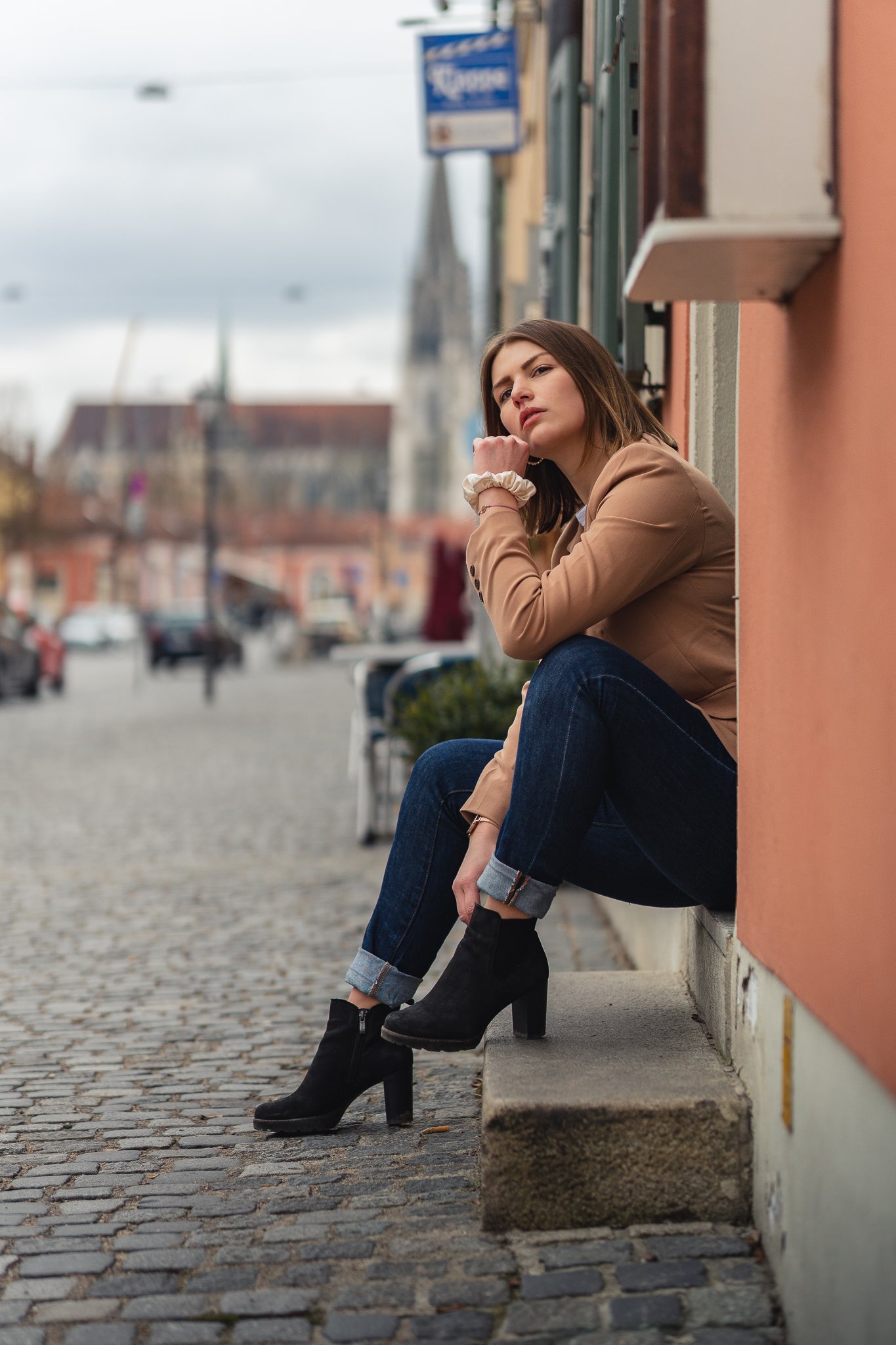 Junge Frau sitzt auf einem Bürgersteig in einer Stadt, trägt schwarze Stiefeletten, dunkelblaue Jeans und einen beigen Mantel, schaut nachdenklich.