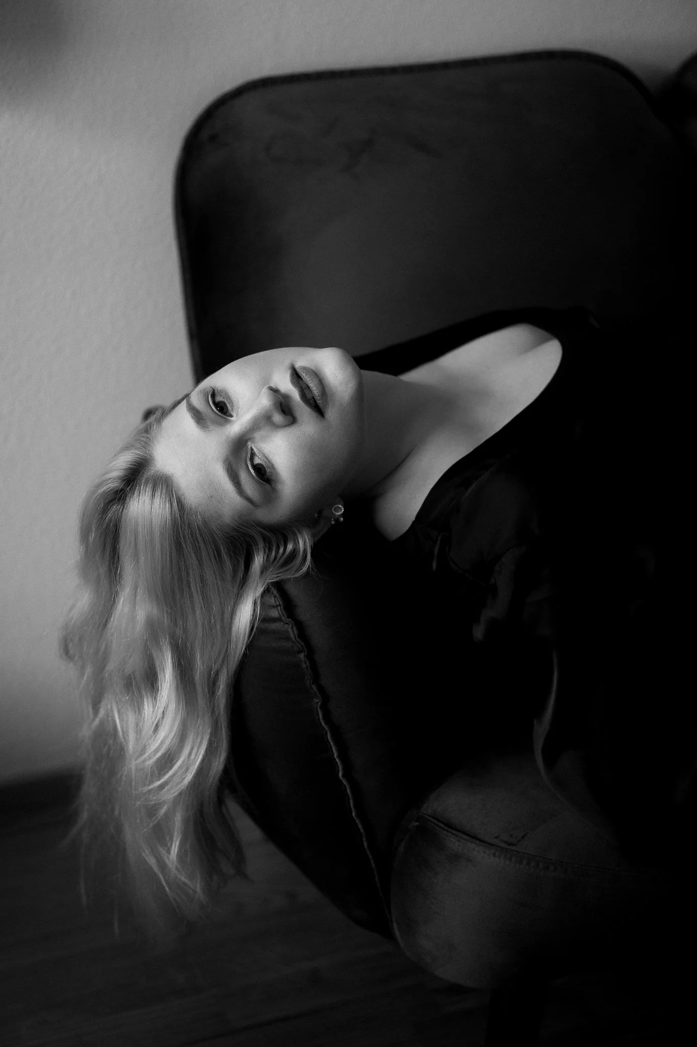 Schwarz-Weiß-Fotografie einer jungen Frau mit langen, gewellten Haaren, die auf einem Stuhl liegt und direkt in die Kamera schaut.