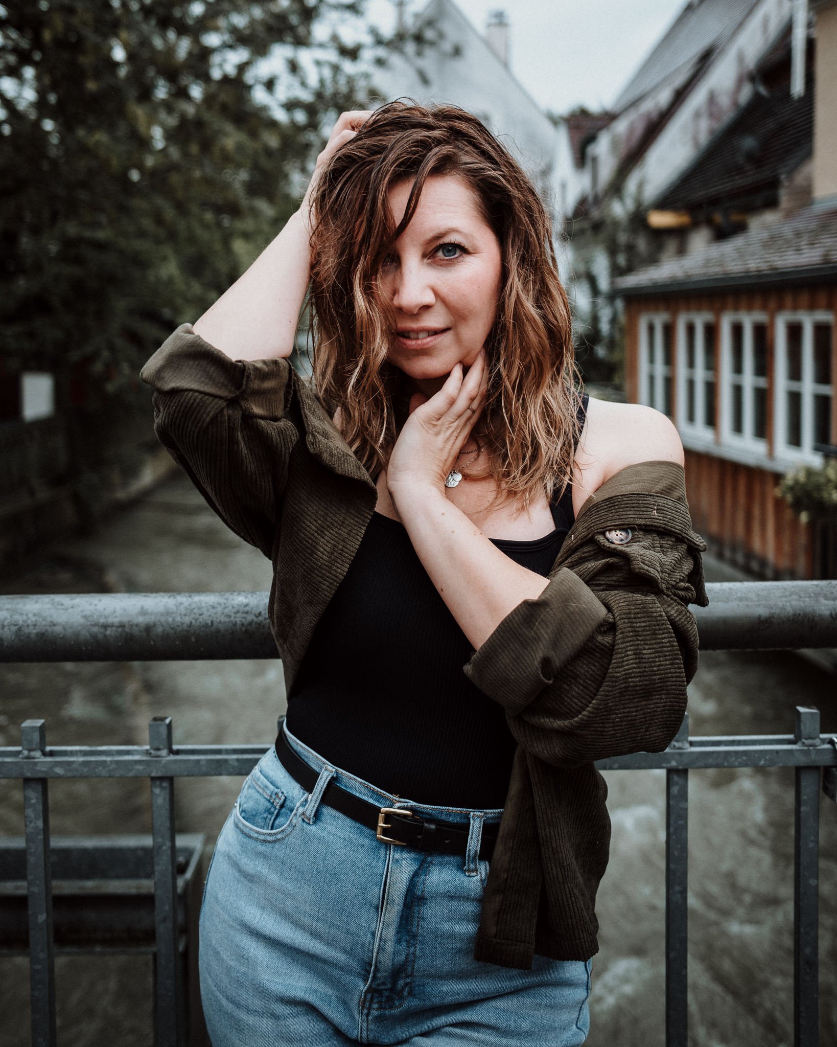 Junge Frau mit braunen, lockigen Haaren, die auf einer Brücke steht, mit grauen und weißen Häusern im Hintergrund, trägt ein schwarzes Top, eine grüne Jacke und blaue Jeans.