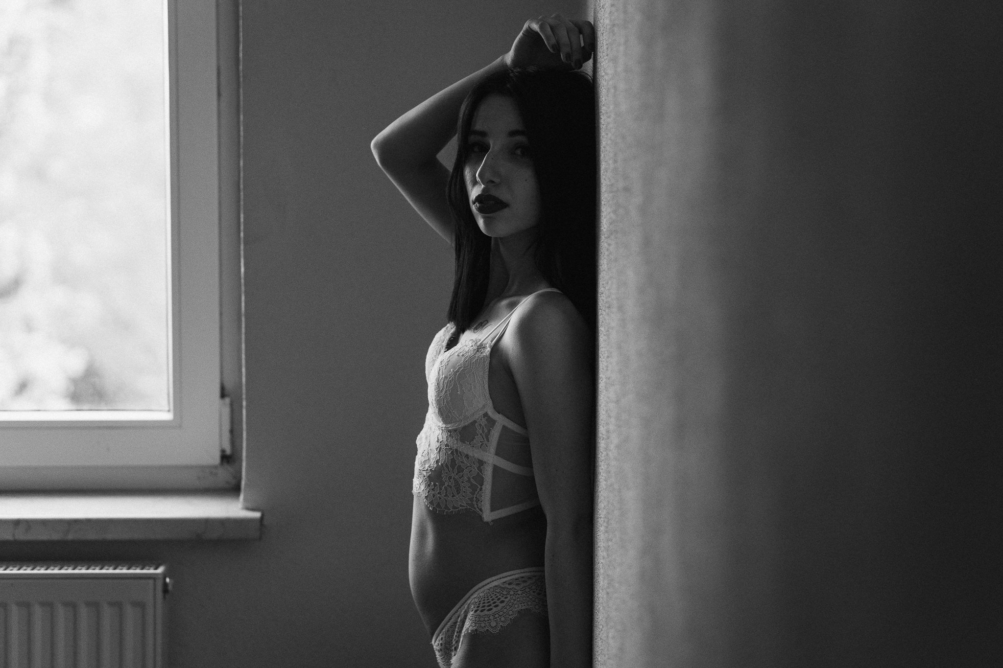 Schwarz-Weiß-Foto einer Frau in Spitze-Breisou, die an einer Wand steht und durch ein Fenster blickt.
