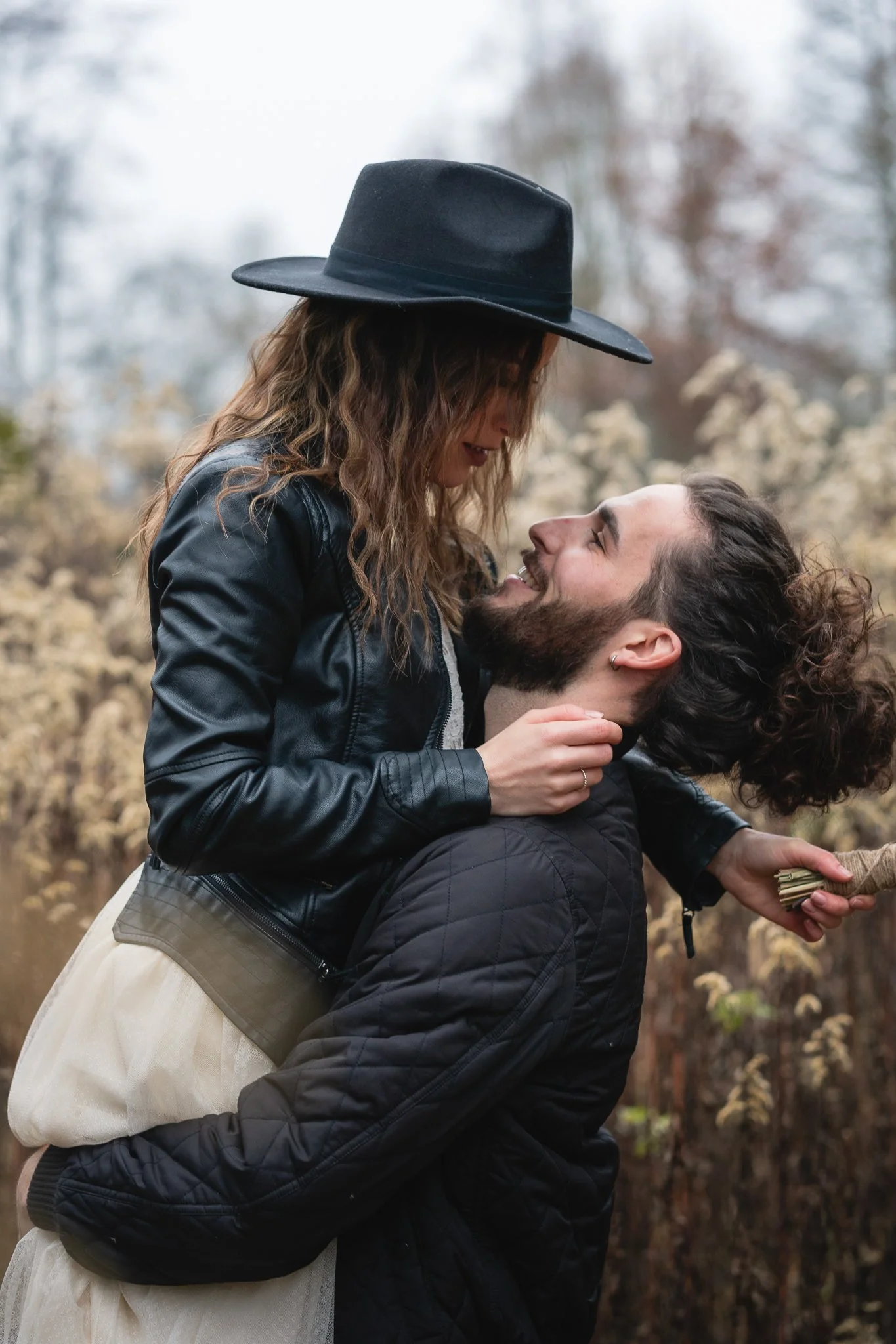 Ein Paar umarmt und schaut sich verliebt an im Freien, die Frau trägt einen schwarzen Hut und Lederjacke, der Mann trägt eine schwarze Jacke.