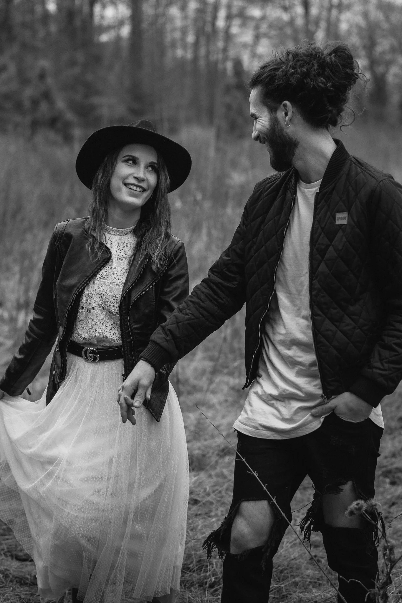 Zwei junge Menschen halten Händchen, lachen und schauen sich liebevoll an, während sie im Freien in einer Wiese mit Bäumen im Hintergrund stehen.