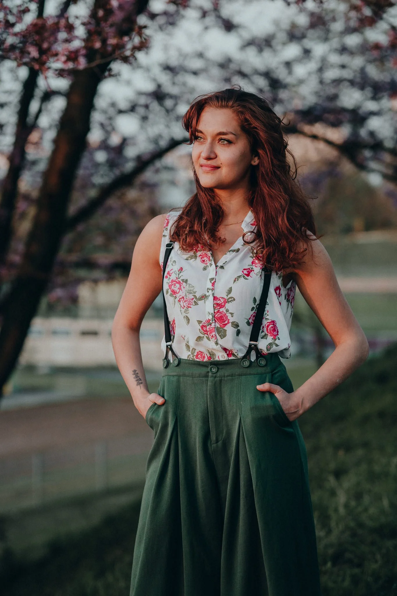 Junge Frau mit roten Locken, gestreiftem Top mit Blumenmuster, grünen hohen Hosen, im Park mit blühenden Bäumen im Hintergrund.