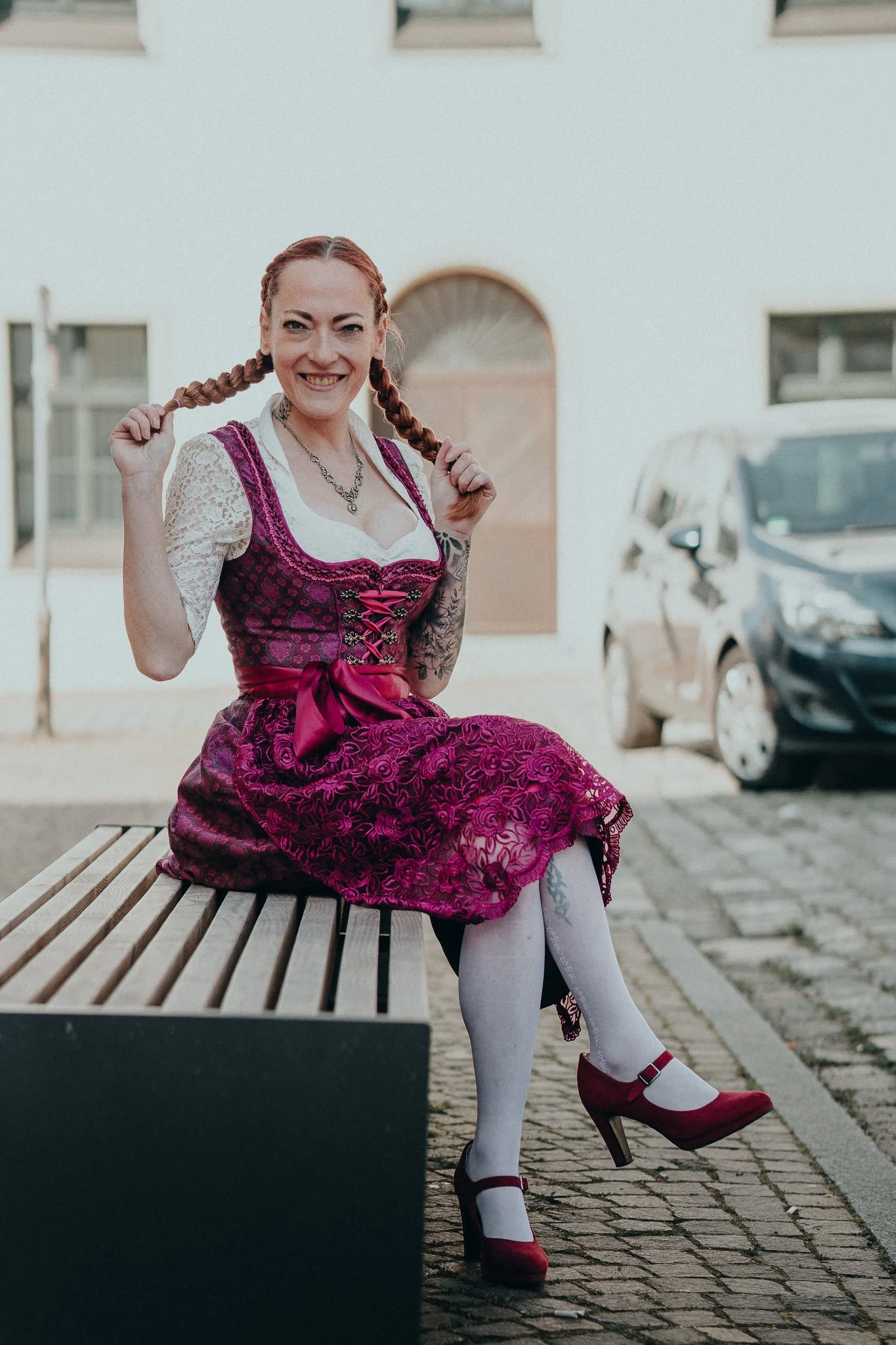 Eine Frau in traditionellem bayerischem Dirndl sitzt auf einer Bank im Freien, hält ihre Zöpfe und lächelt in die Kamera.