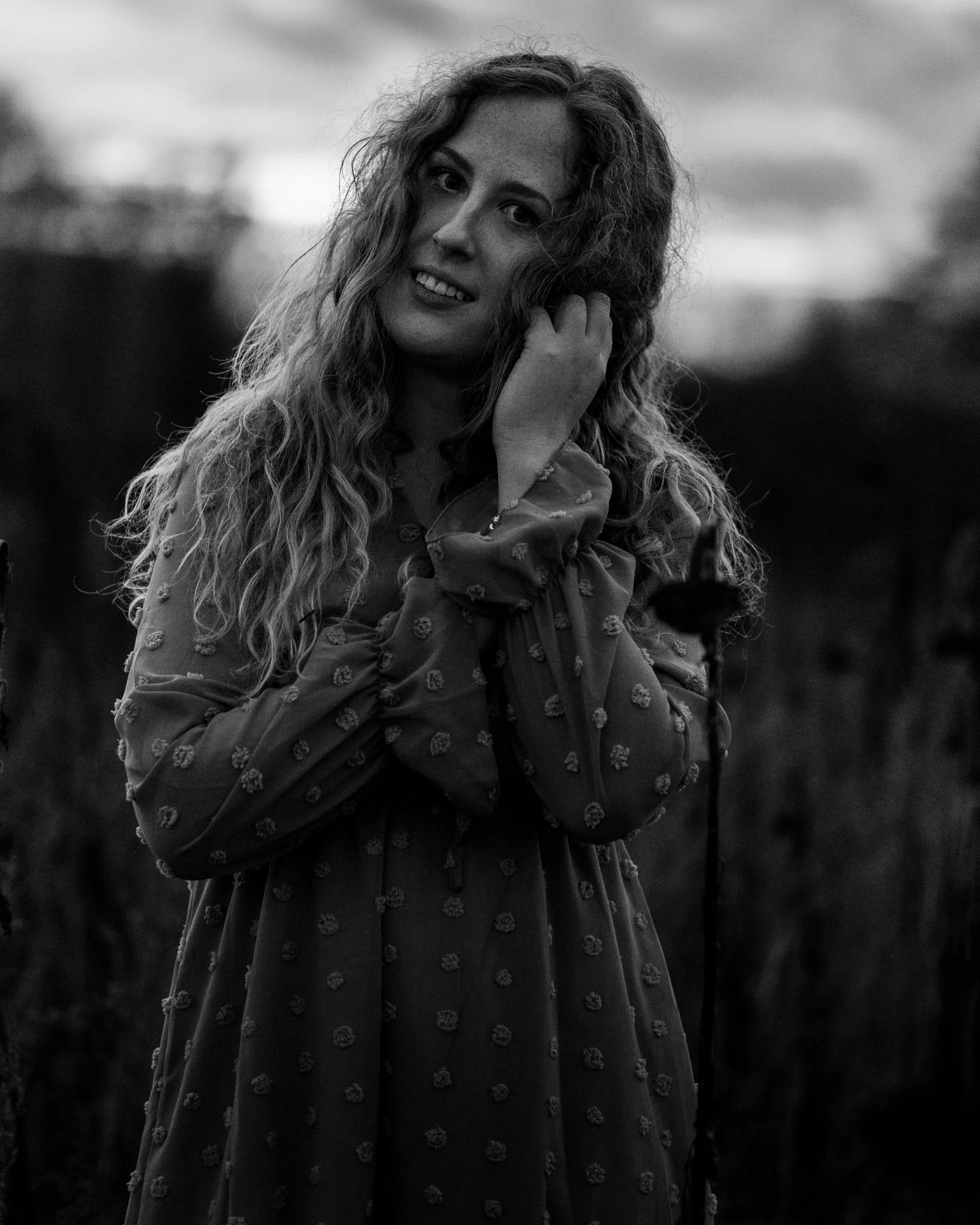 Schwarz-weiß-Foto einer jungen Frau mit lockigem Haar, die eine Hand an ihr Ohr hält, im Freien mit unscharfem Hintergrund bei Dämmerung.