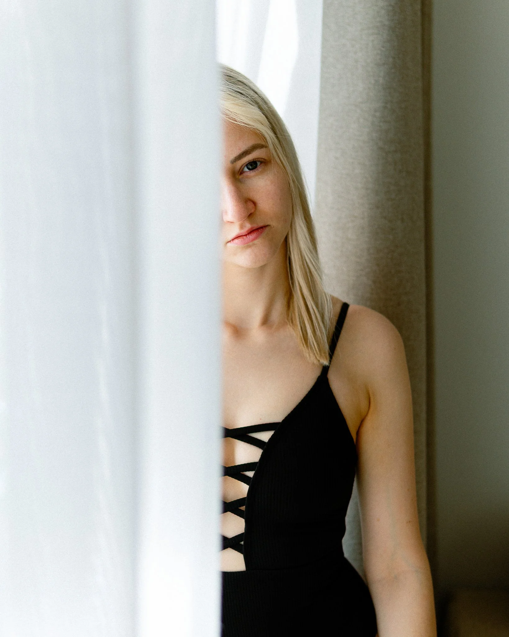 Junge Frau mit blonden Haaren steht neben einem Fenstervorhang und schaut nach vorne.