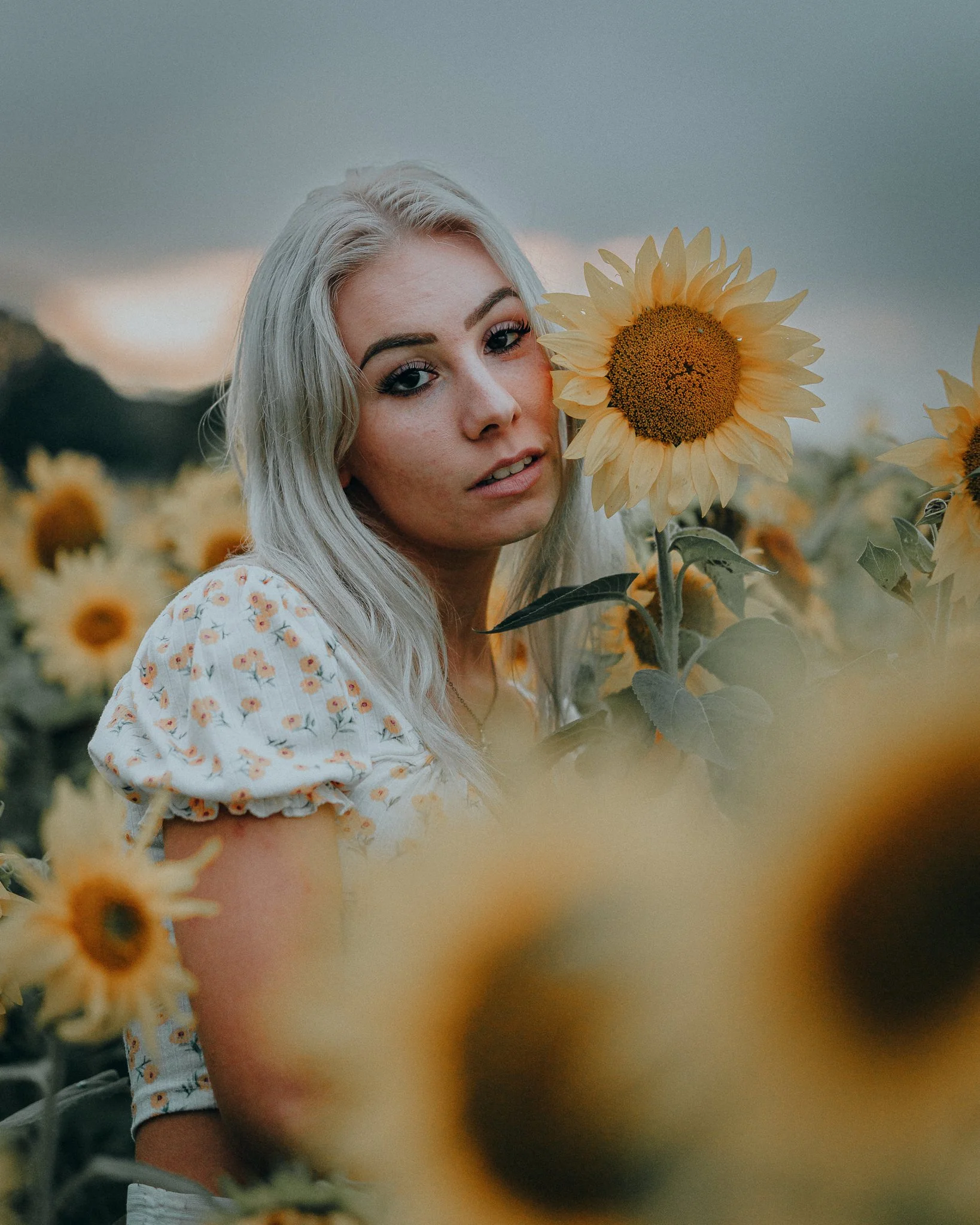 Eine junge Frau mit blonden Haaren und Blümchenmuster-Kleid steht in einem Sonnenblumenfeld, hält eine Sonnenblume nahe an ihr Gesicht, bei bewölktem Himmel.