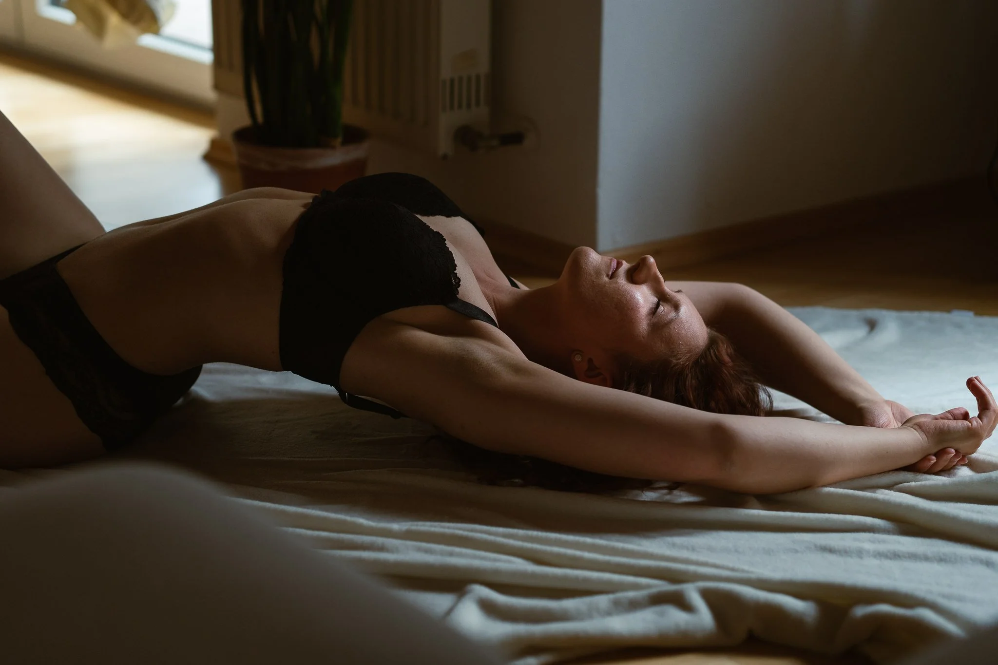 Frau liegt entspannt auf einem Bett, mit ausgestreckten Armen hinter dem Kopf, in einem dunklen Raum, bei Tageslicht