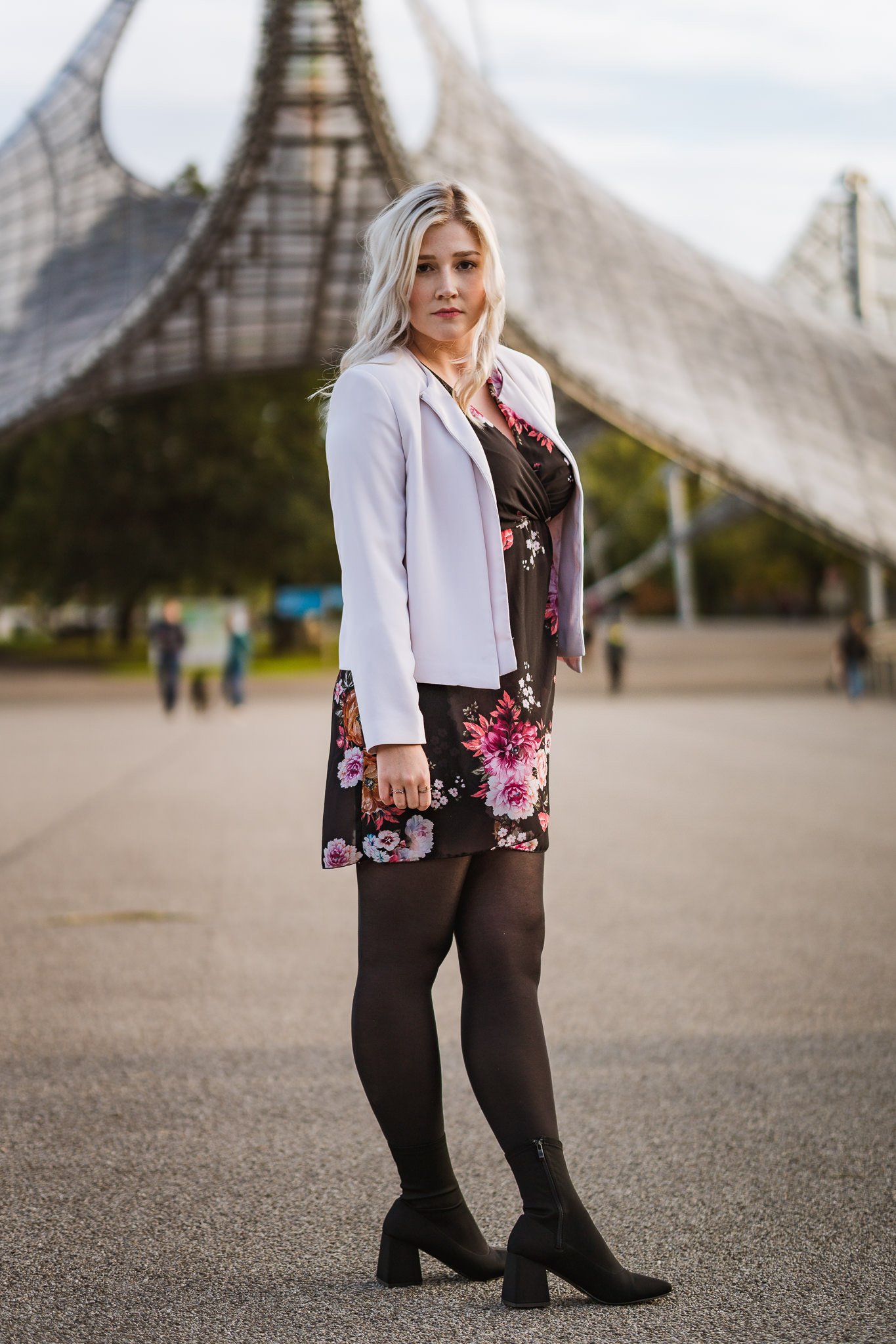 Junge Frau mit schwarzem floralem Kleid, weißer Jacke, schwarzen Strumpfhosen und schwarzen Stiefeln steht auf einer Freifläche vor moderner, wellenförmiger Architektur.
