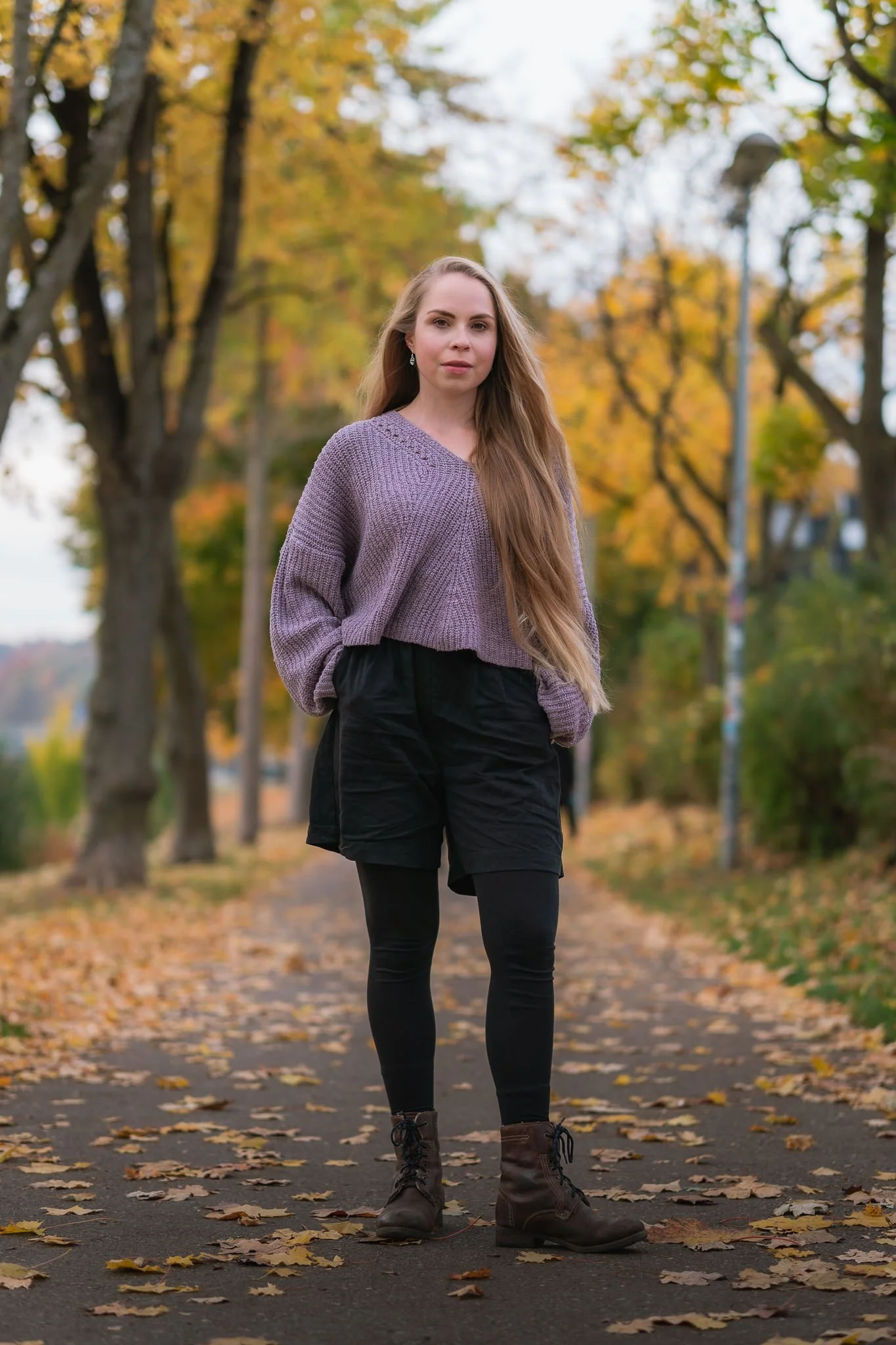 Junge Frau in Herbstjacke und Stiefeln steht auf einem fallenblätterbedeckten Weg in einem Park mit bunten Herbstbäumen im Hintergrund.