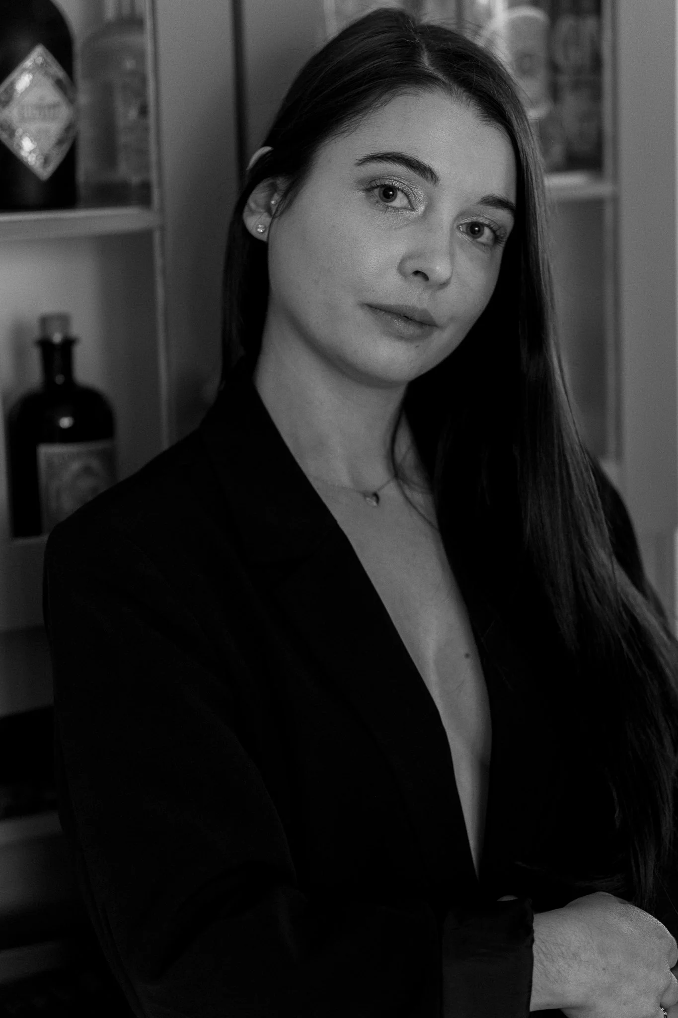Schwarz-Weiß-Foto einer jungen Frau mit langen, glatten Haaren, die an einer Bar oder einem Regal steht, mit Flaschen im Hintergrund.