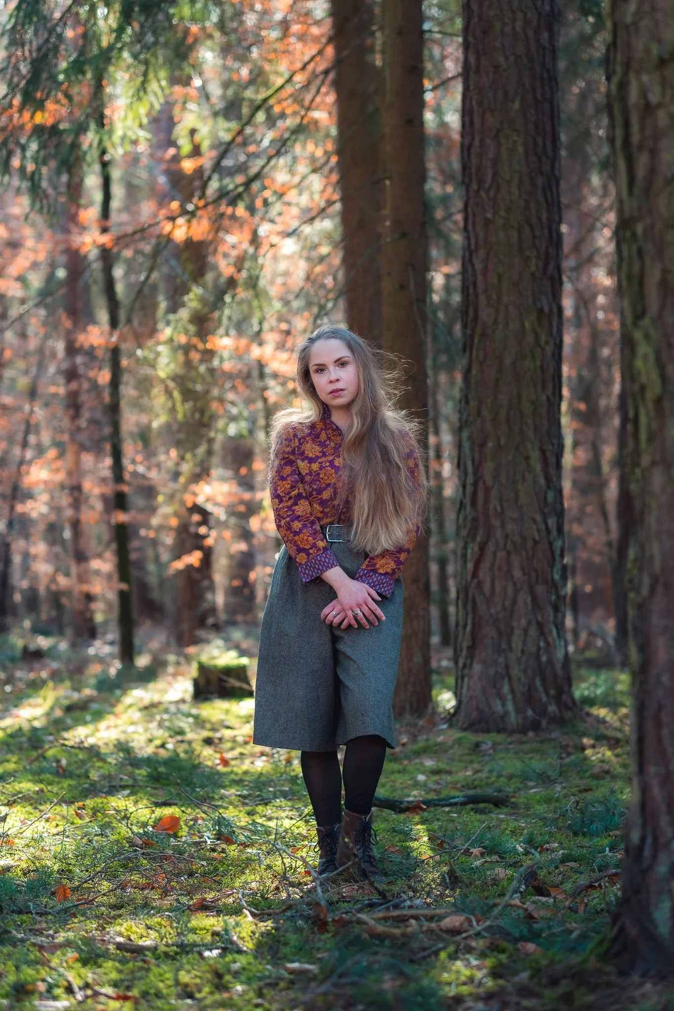 Eine junge Frau mit langen, welligen Haaren steht in einem Herbstwald mit herbstlichen Blättern und Bäumen im Hintergrund.