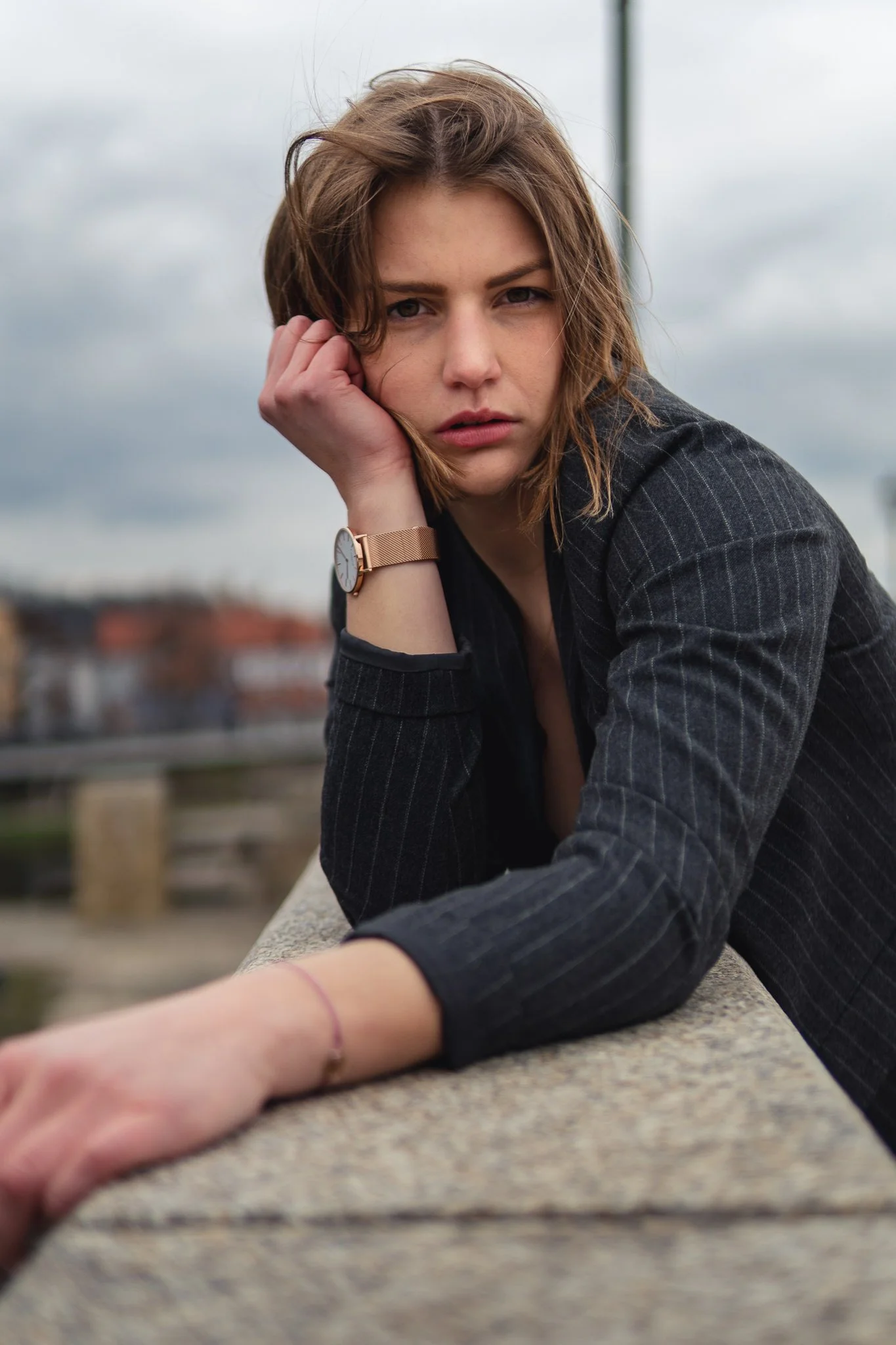 Junge Frau mit kurzen braunen Haaren, trägt schwarze Jacke und einen Watch, lehnt auf einem Geländer im Freien bei bewölktem Himmel an.