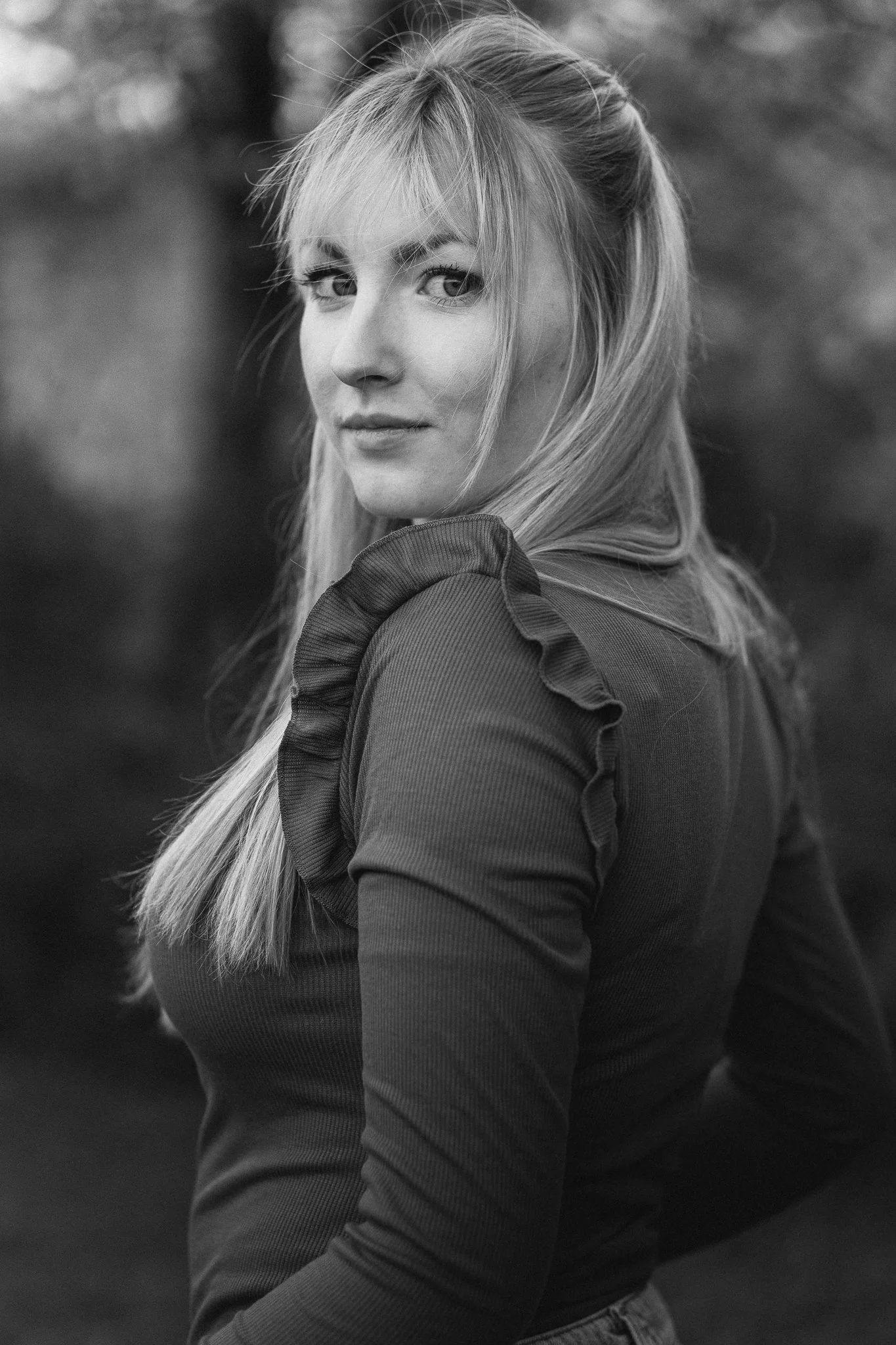 Schwarz-weiß Porträt einer jungen Frau mit langen, geraden Haaren, die ein schwarzes Oberteil mit Rüschen an den Schultern trägt, im Freien mit unscharfem Hintergrund.