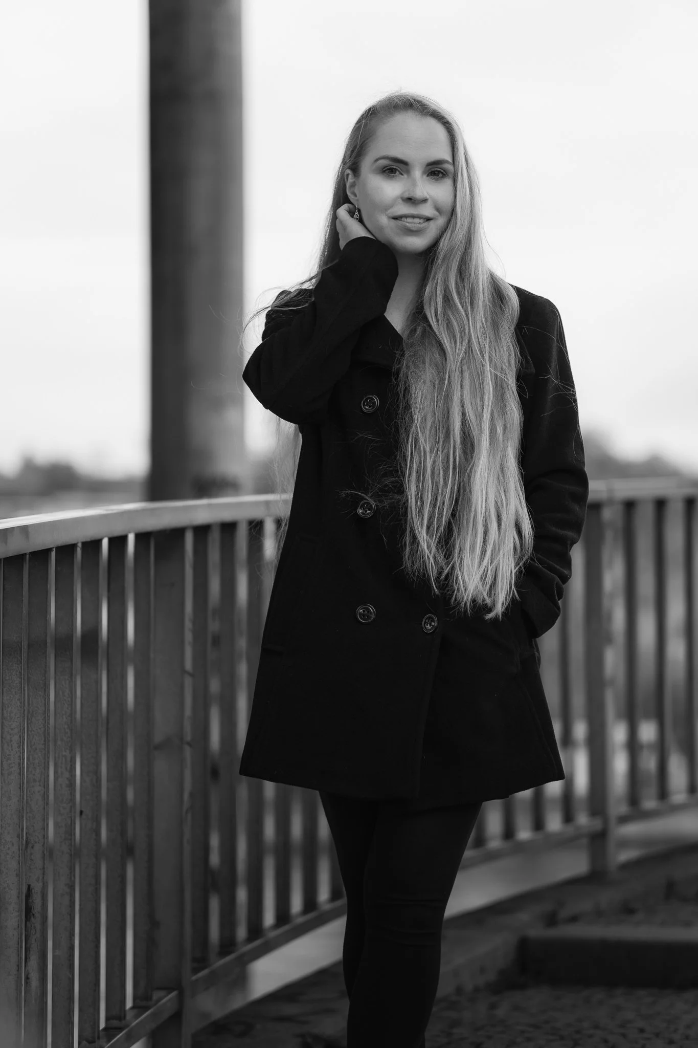 Schwarz-weiß-Foto einer jungen Frau mit langen, welligen Haaren, die eine schwarze Jacke trägt, auf einer Brücke stehend, lächelnd, mit einer Hand am Hals, im Hintergrund ein Himmel und eine ländliche Landschaft.