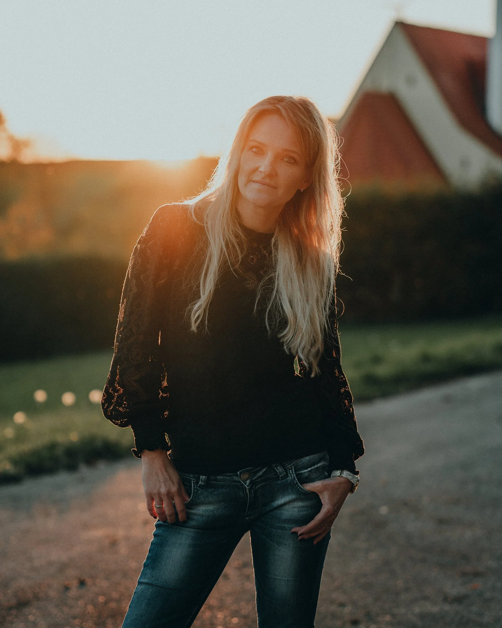 Frau steht bei Sonnenuntergang im Freien, trägt schwarze Bluse und Jeans, Sonnenstrahlen beleuchten hinter ihr den Himmel.