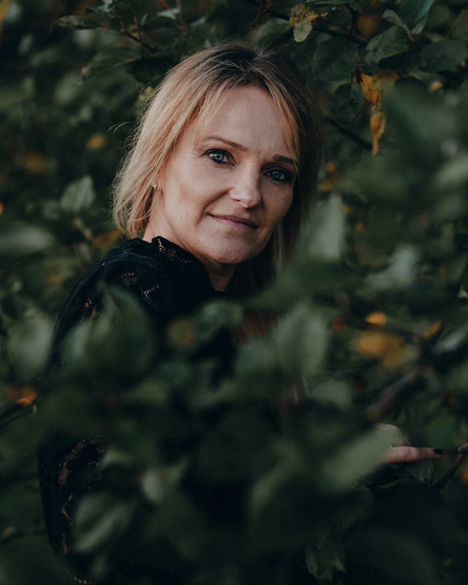 Nahaufnahme einer Frau mit Blondhaar, die durch grüne Blätter schaut.