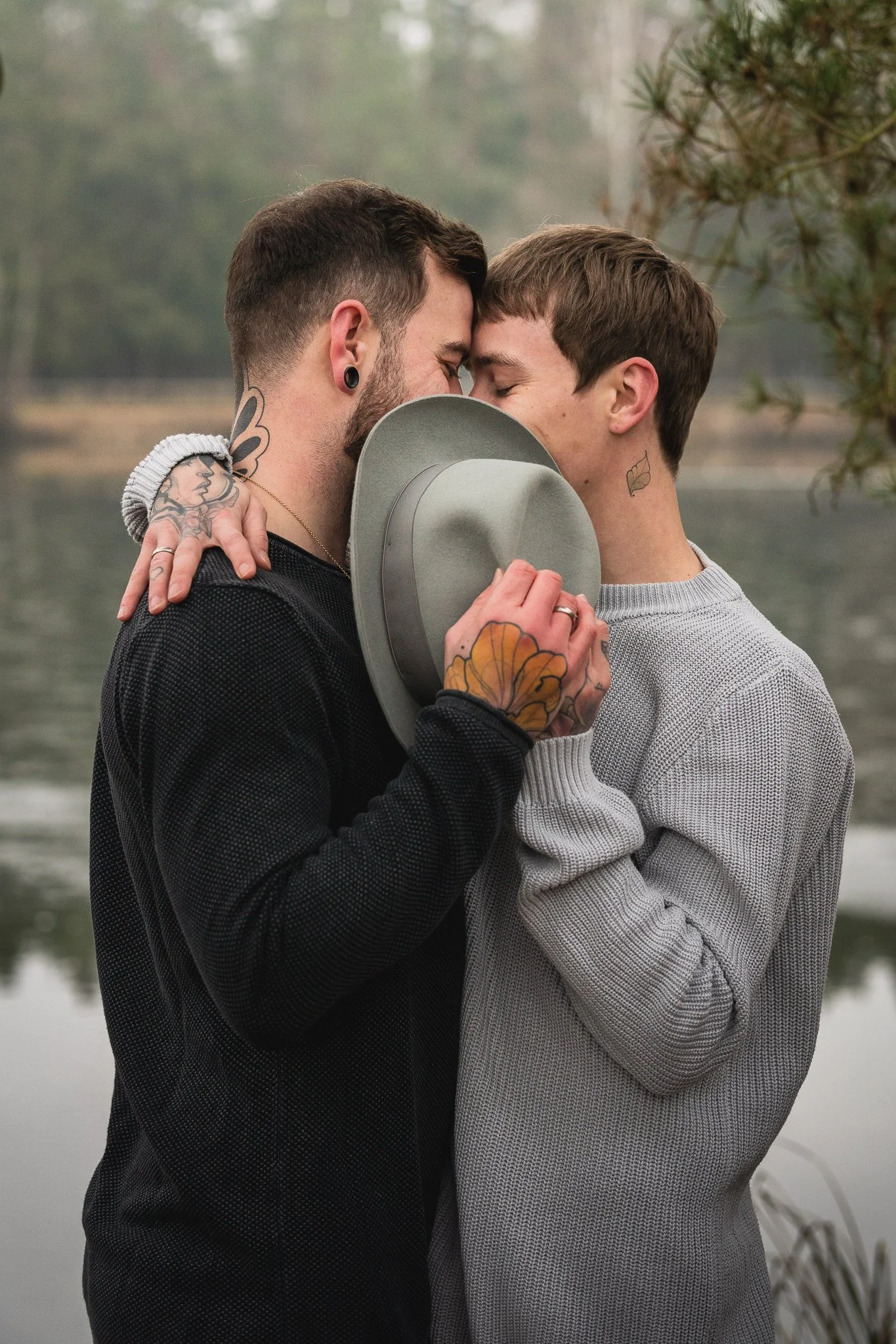 Zwei Männer umarmen sich verliebt am See, halten einen Hut zwischen ihren Stirnflächen, Sonnenbrille auf dem Hut, im Hintergrund Bäume und Wasser.