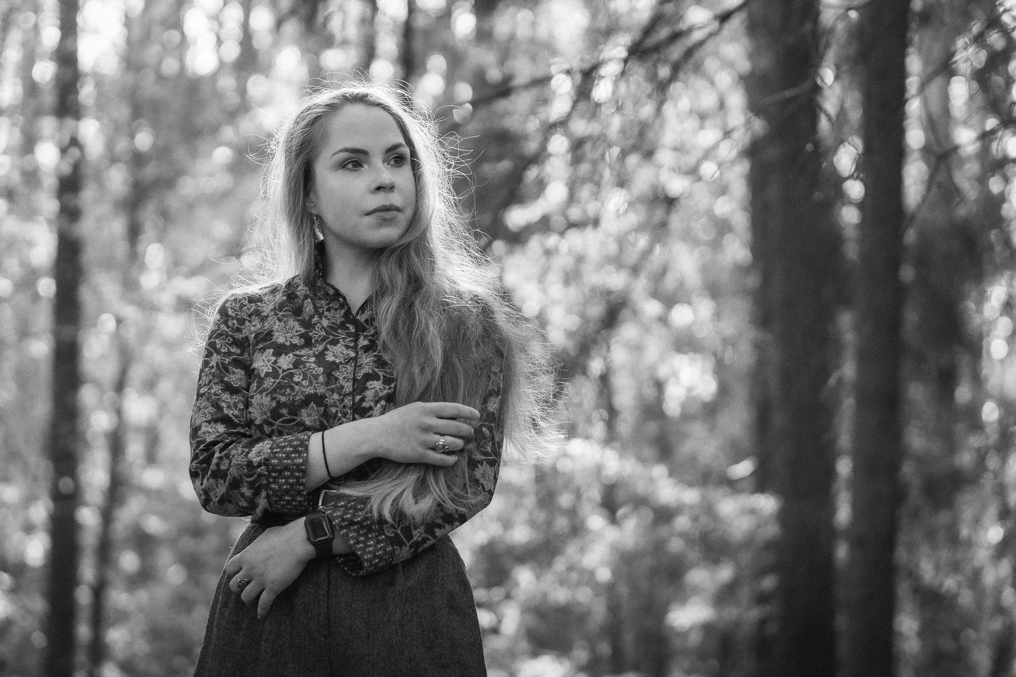Junge Frau mit langen Haaren in einem Wald, trägt eine blumenverzierte Bluse und einen Rock, schaut nach rechts, in Schwarzweiß aufgenommen.