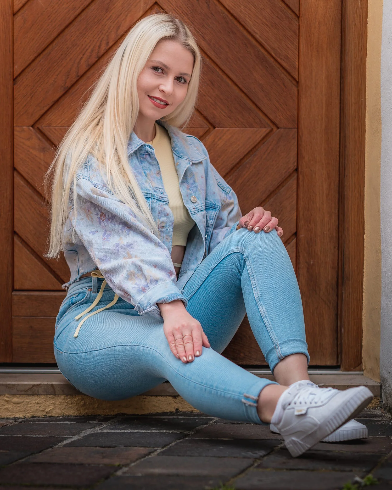 Junge Frau mit blonden Haaren, blauer Jeans und Jacke sitzt vor einer Holztür.