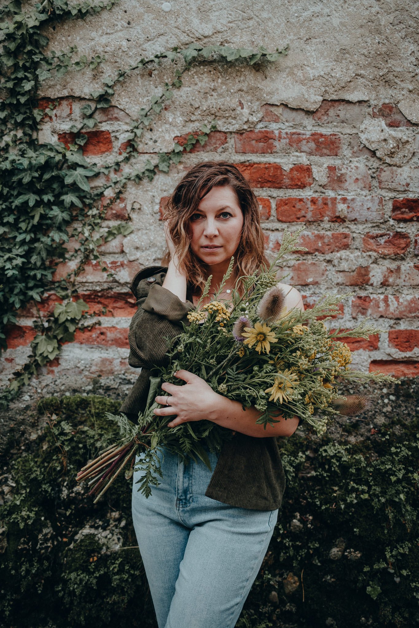 Eine junge Frau mit lockigen braunen Haaren hält einen großen Blumenstrauß mit wilden Blumen vor einer alten Ziegelmauer, umgeben von grünem Bewuchs.