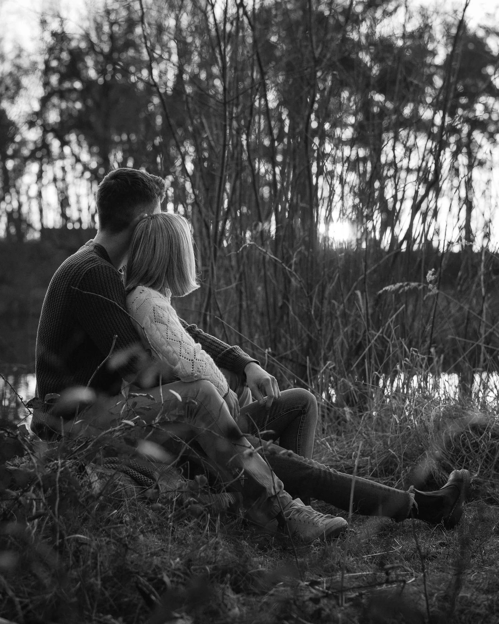 Ein Mann und ein Kind sitzen nebeneinander an einem Fluss im Wald bei Sonnenuntergang, die Szenerie ist in Schwarzweiß fotographiert.