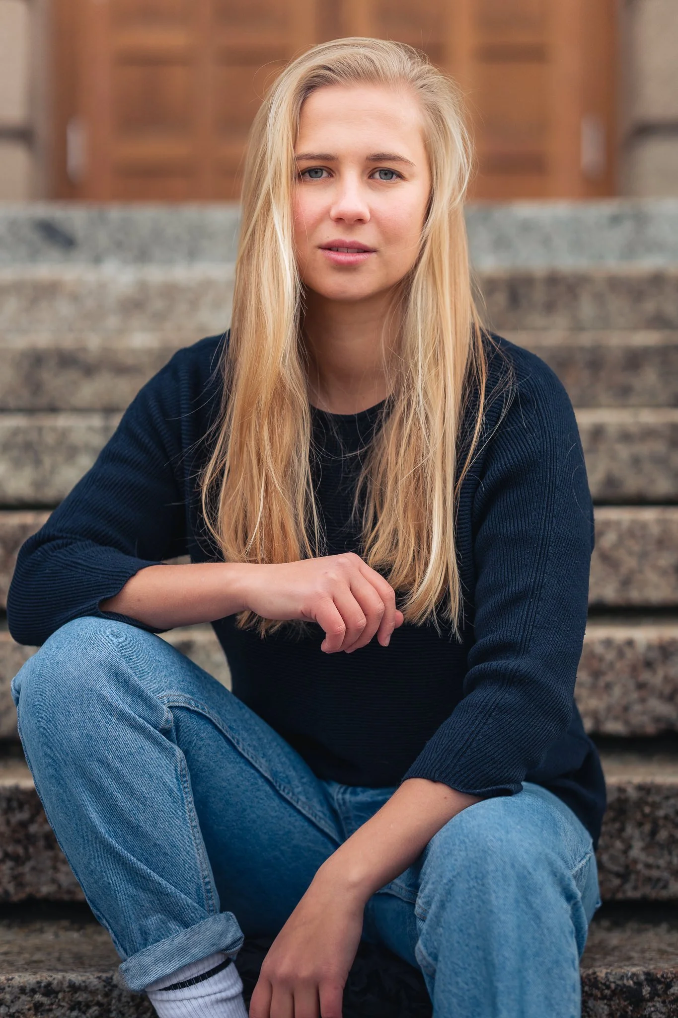 Junge Frau mit langem blondem Haar in dunklem Pullover und blauen Jeans sitzt auf Steintreppe vor einer Holztür.
