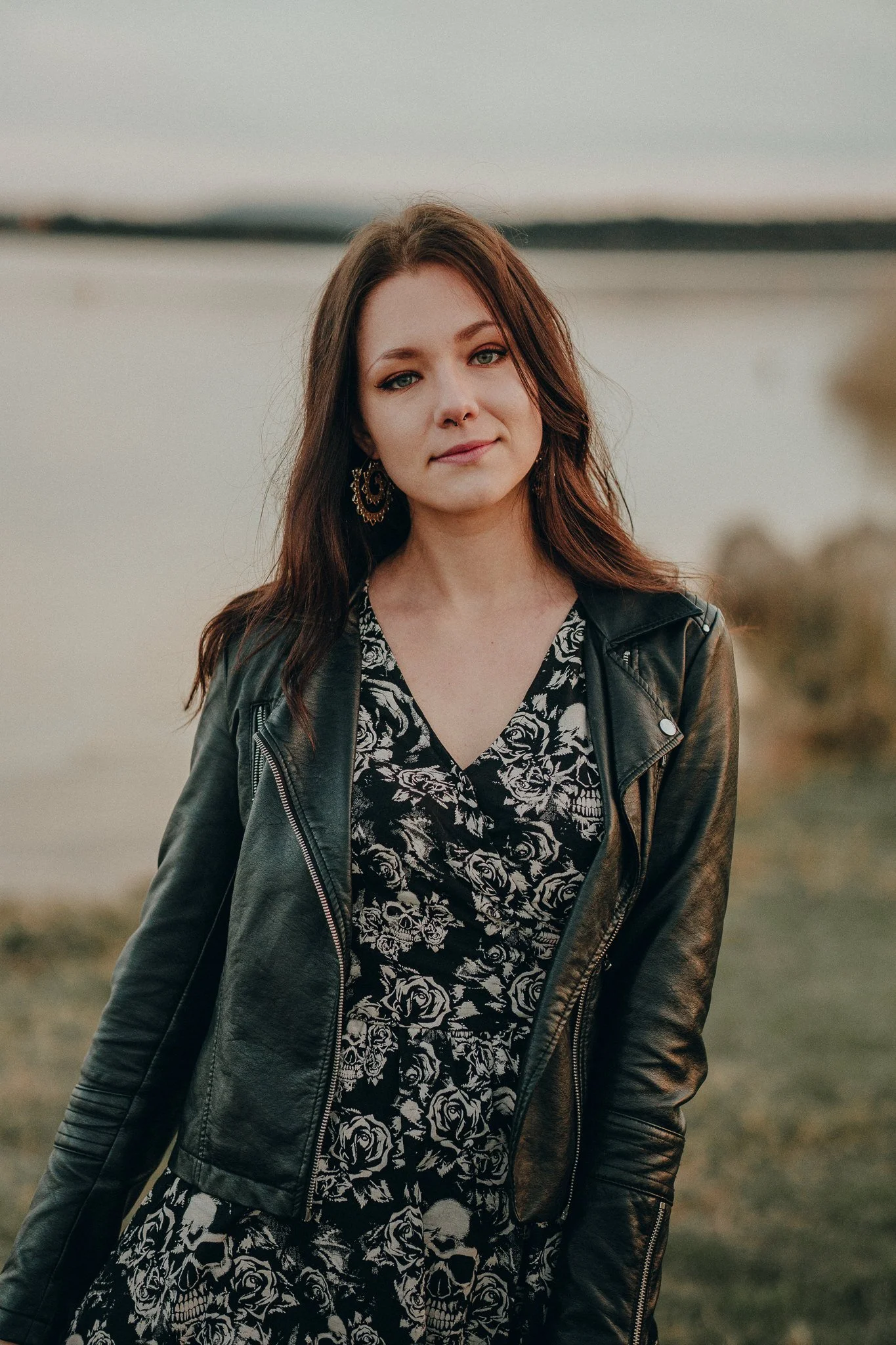Eine junge Frau mit braunen Haaren, die eine schwarze Lederjacke und ein schwarzes Kleid mit weißen Rosenmustern trägt, steht vor einem Wasserhintergrund.