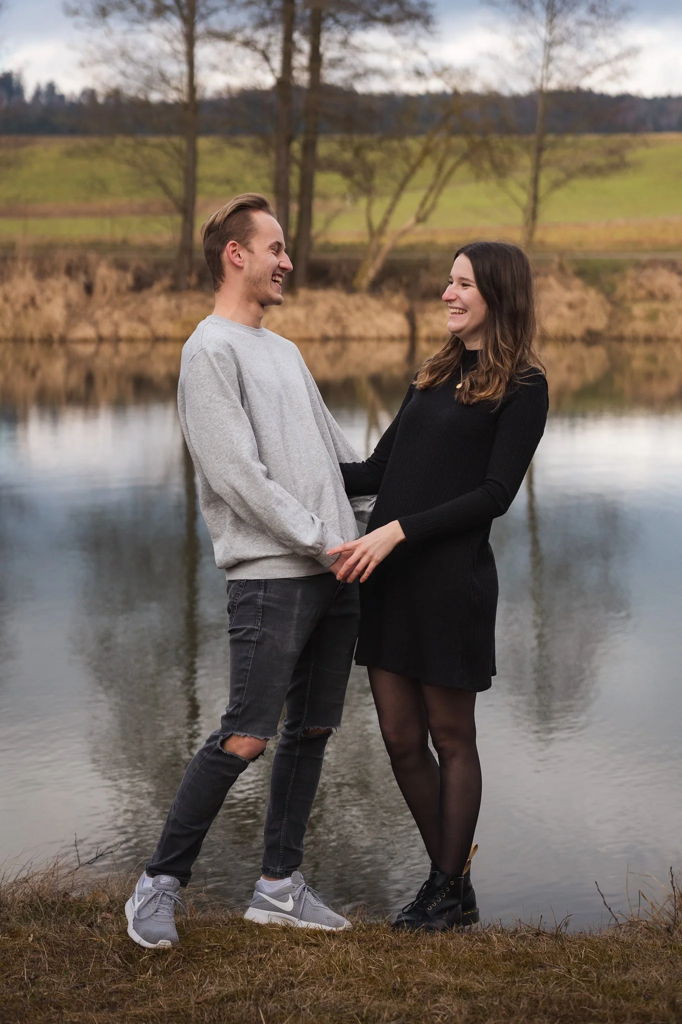 Ein lachendes Paar steht am Flussufer, die Frau hält die Hand des Mannes, im Hintergrund Bäume und Wasser, herbstliche Umgebung.