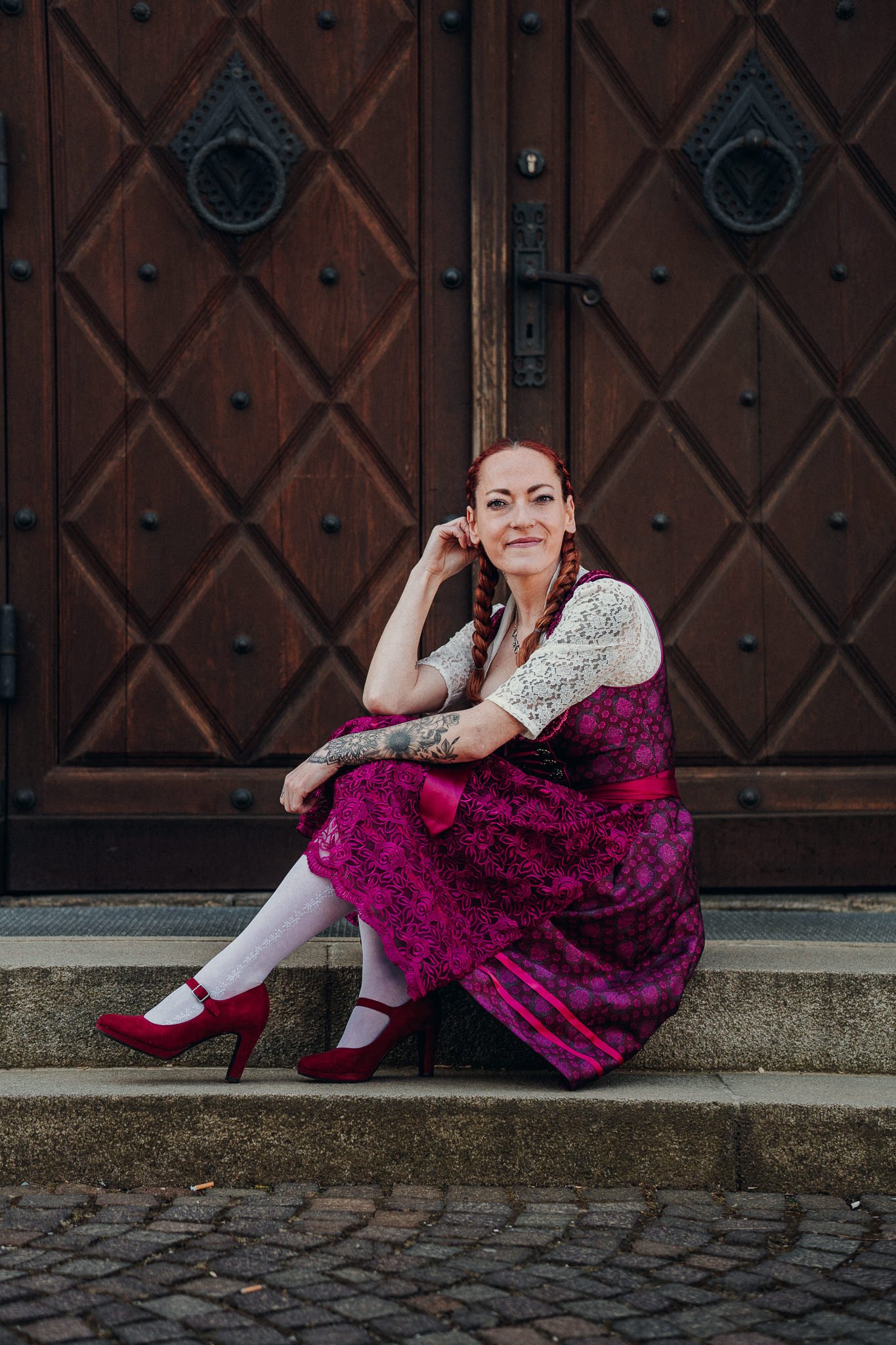 Frau mit roten Zöpfen in einem traditionellen Dirndl sitzt auf den Stufen vor einer großen Holztür.