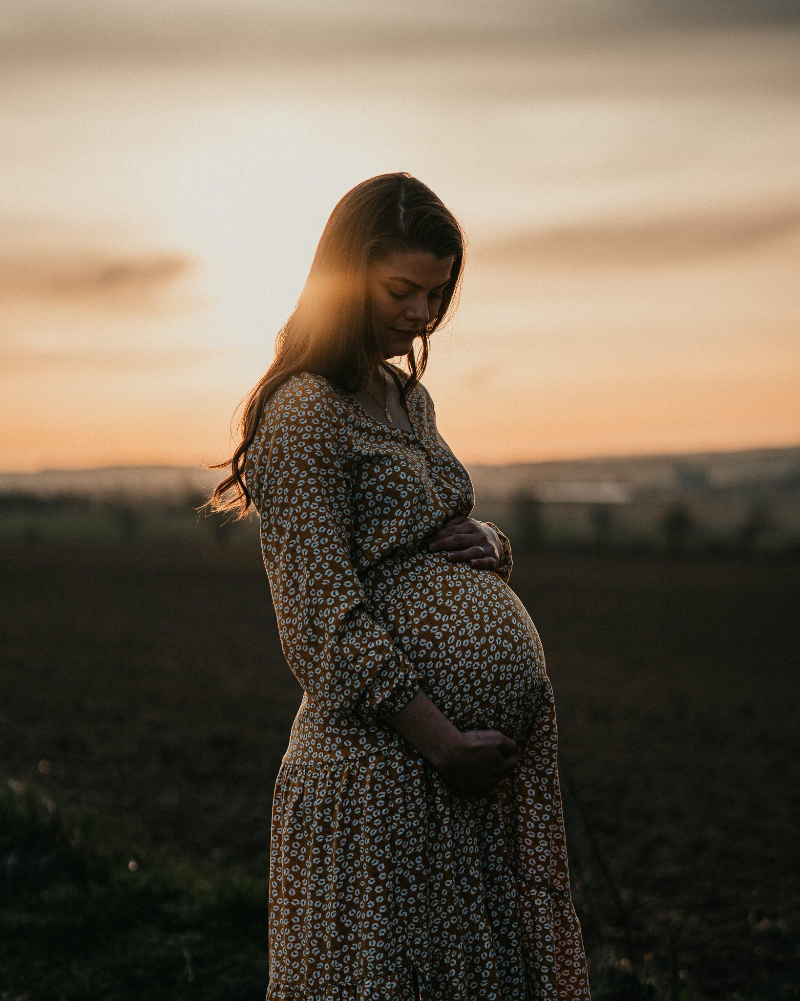 Eine schwangere Frau mit langen braunen Haaren steht im Freien während des Sonnenuntergangs, trägt ein langes Kleid mit braun-weißen Leopardmuster, hält ihre Hände auf den Bauch und blickt nach unten.