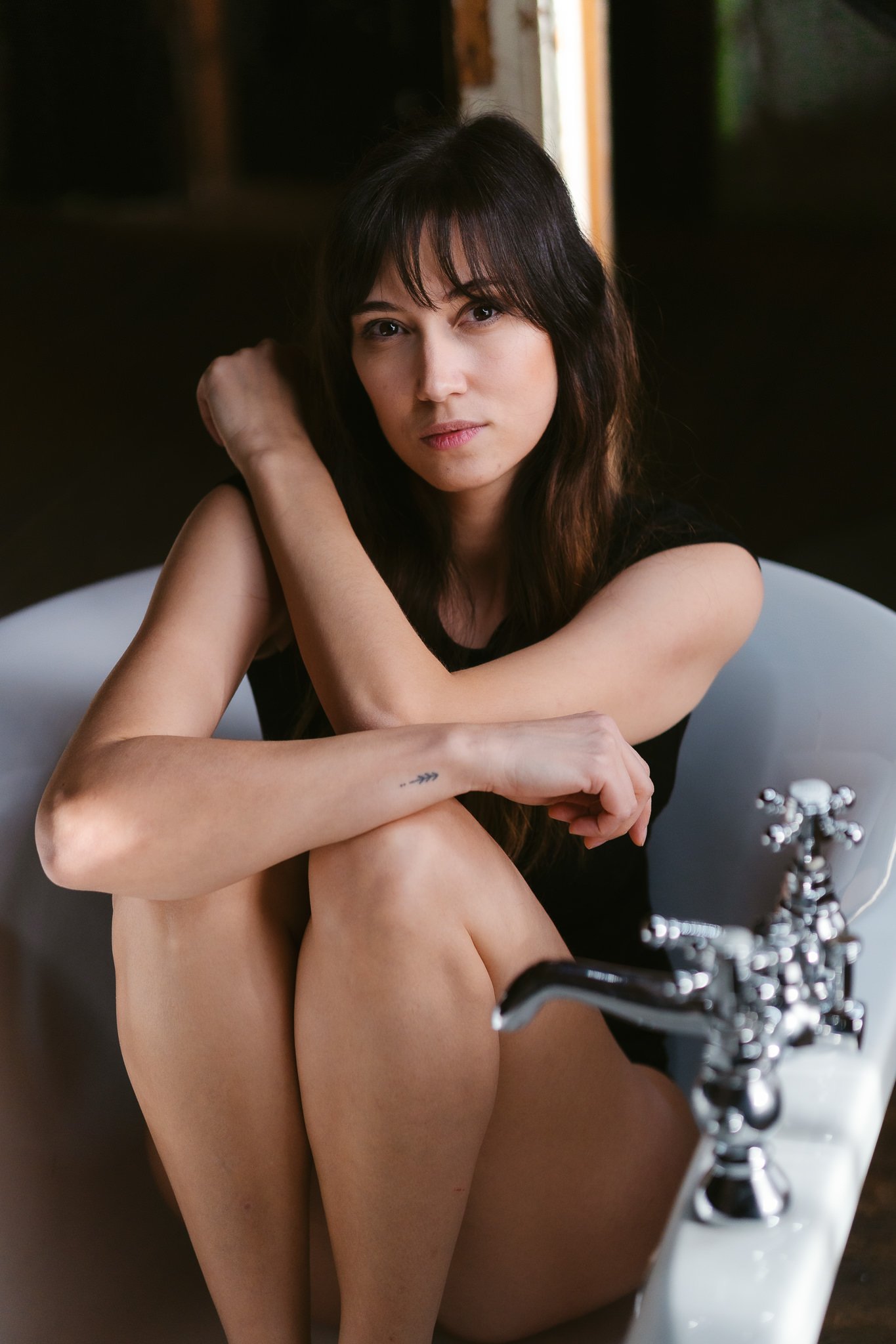 Junge Frau mit dunklen Haaren sitzt in einer Badewanne, umgeben von dunklem Raum, schaut direkt in die Kamera.
