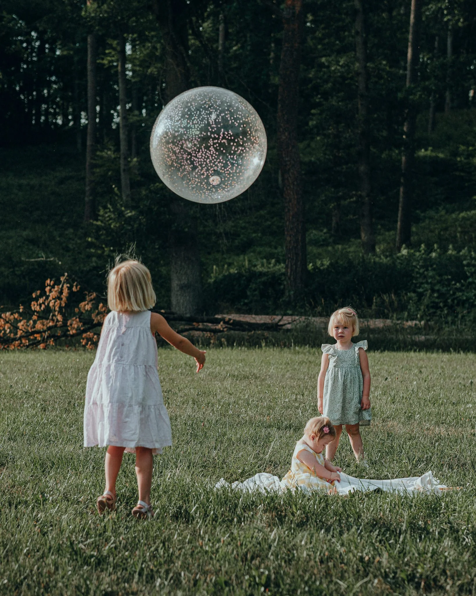 Drei kleine Mädchen spielen im Freien auf einer Wiese, eines wirft einen Luftballon, während die anderen beiden zuschauen.