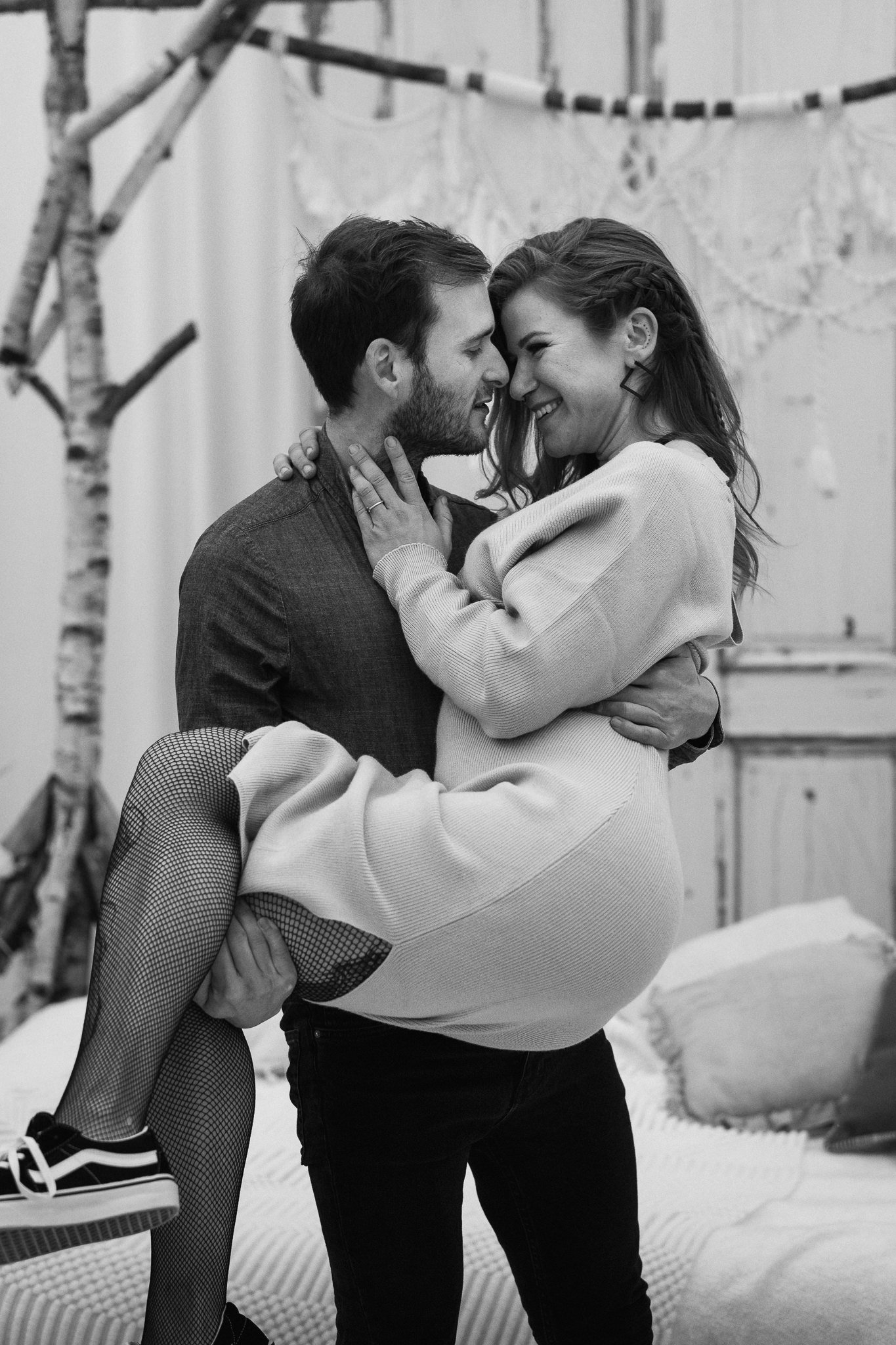 Ein Mann trägt eine Frau in seinen Armen, beide lachen und berühren sich liebevoll in einem Raum mit dekorativem Hintergrund.