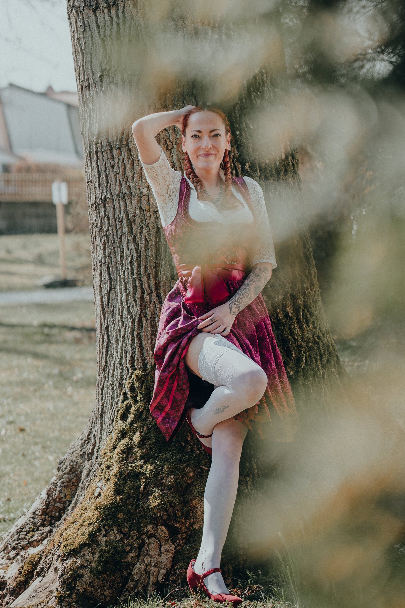 Eine Frau in einem Dirndl mit weißen Strümpfen und roten Schuhen lehnt an einem Baum im Freien, umgeben von Blättern.