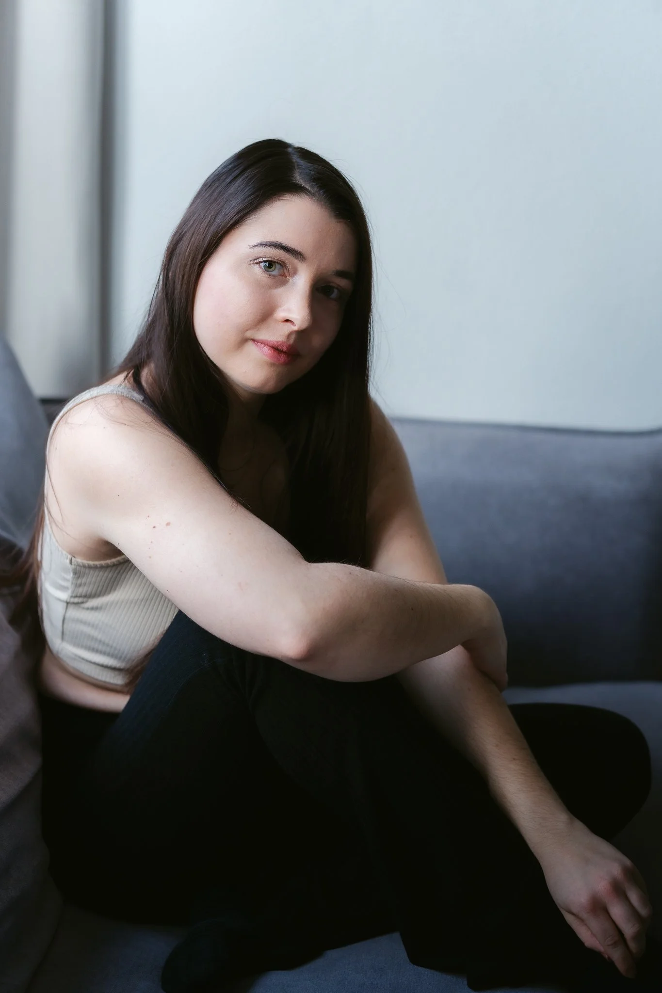 Junge Frau mit langen braunen Haaren, sitzend auf einem grauen Sofa, trägt ein beiges Top und schwarze Hosen, blickt in die Kamera.