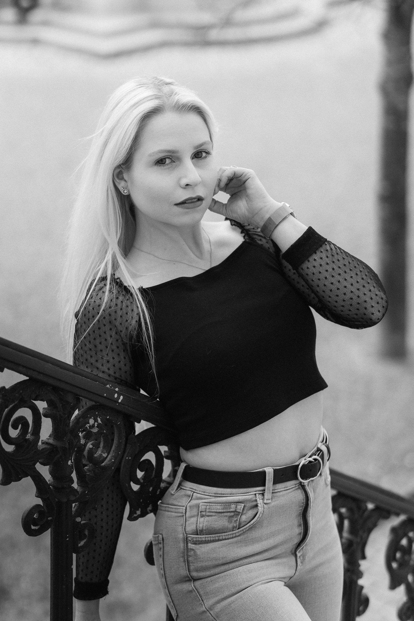 Schwarz-Weiß-Foto einer jungen Frau mit langen, hellen Haaren, die vor einem Zaun steht und in die Kamera schaut, mit einer Hand an ihrem Gesicht, trägt eine schwarze Bluse mit transparenten, gepunkteten Ärmeln und eine Jeans.