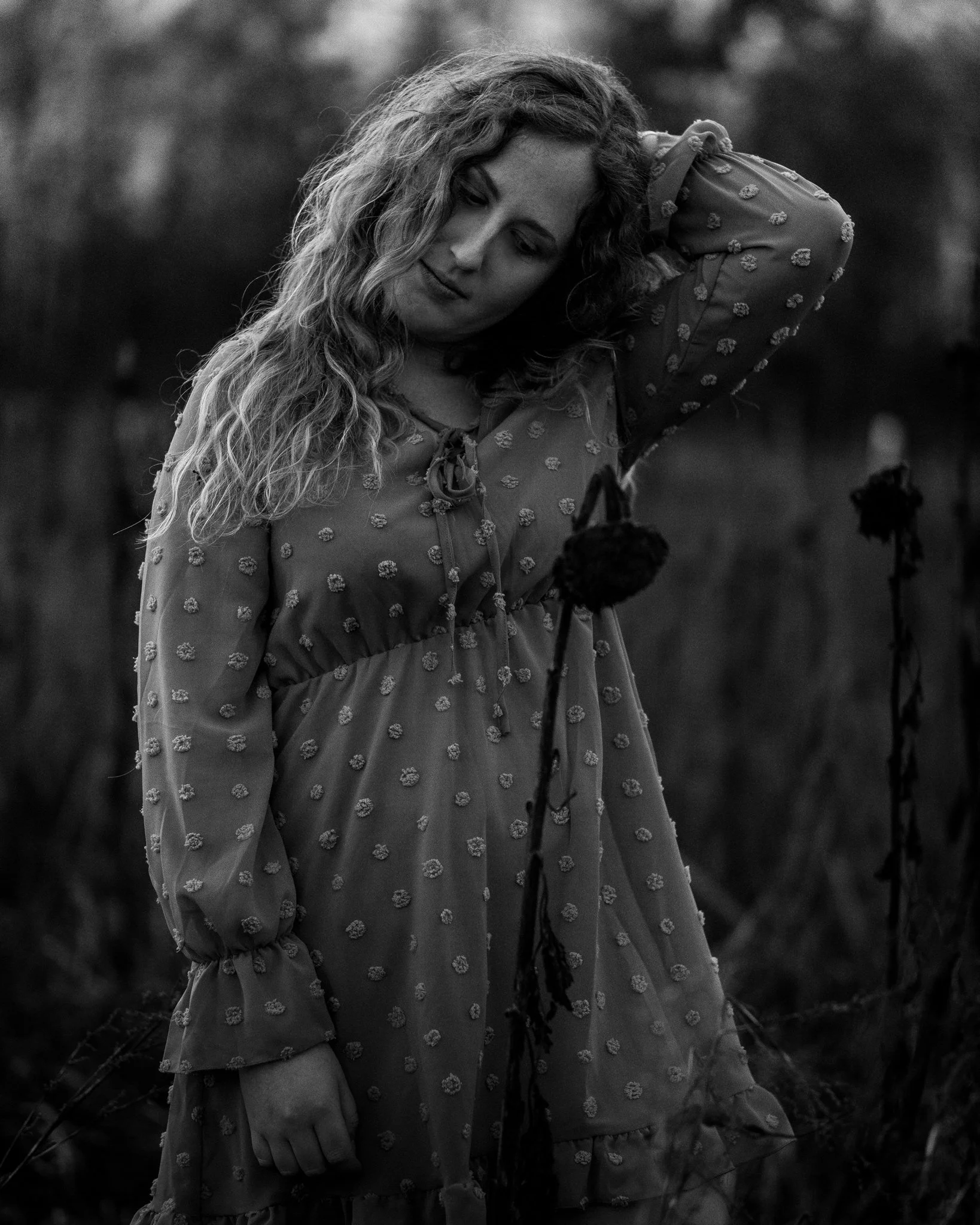 Schwarz-weiß Foto einer jungen Frau mit langen, lockigen Haaren in einem langen Kleid, die auf einem Feld steht und nachdenklich aussieht.
